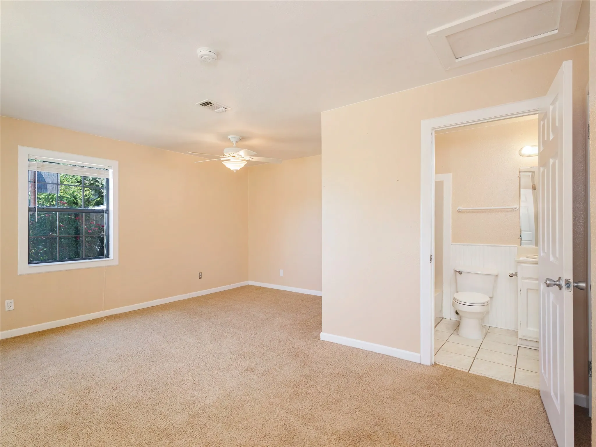 Carpeted room with a ceiling fan, a window, and a white interior door opening to a bathroom with tile flooring, a toilet, and a vanity