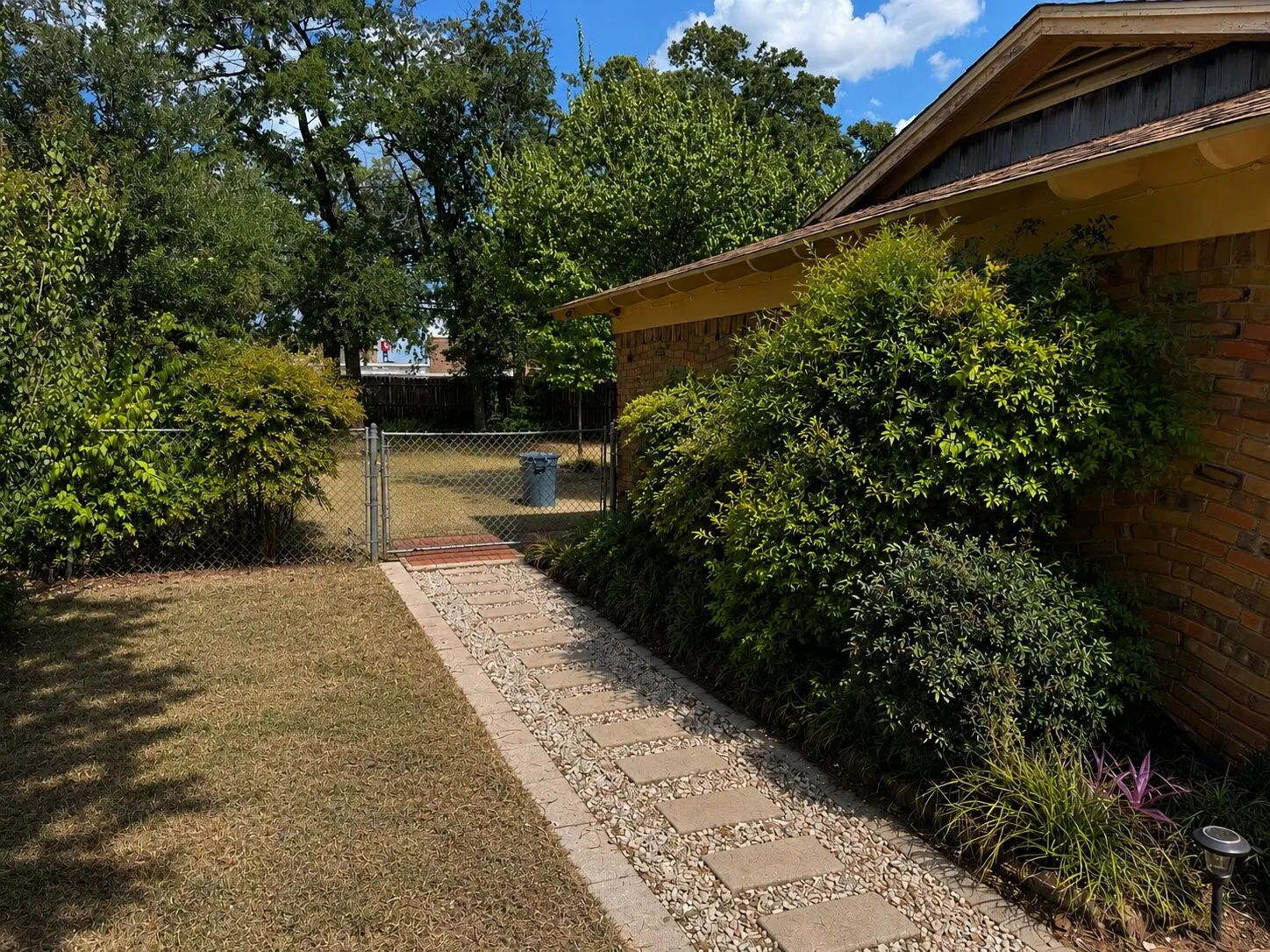 Stone paver pathway with gravel accent, extensive mature landscaping, brick exterior, and a chain-link fence