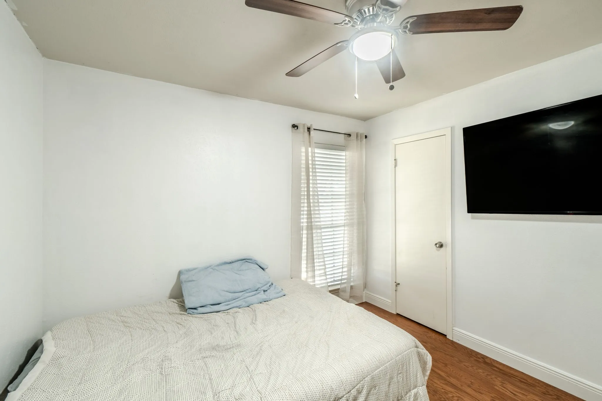 Hardwood-style flooring, ceiling fan with integrated lighting, window with blinds, wall-mounted television, and a white interior door
