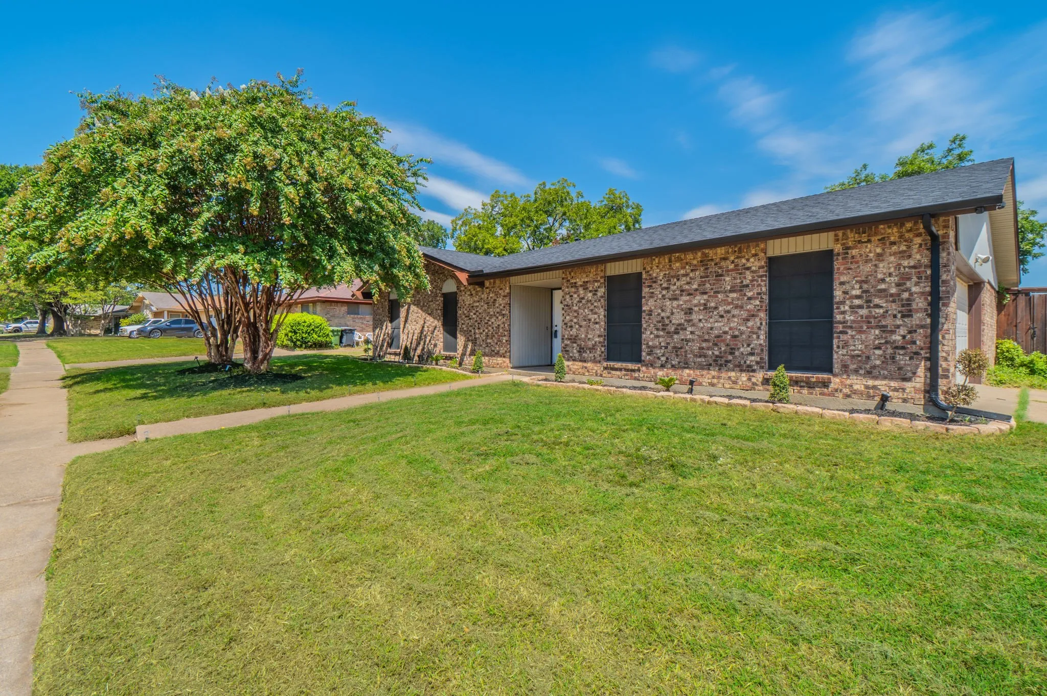 Brick exterior with dark roof shingles, mature tree, established lawn, and concrete sidewalk