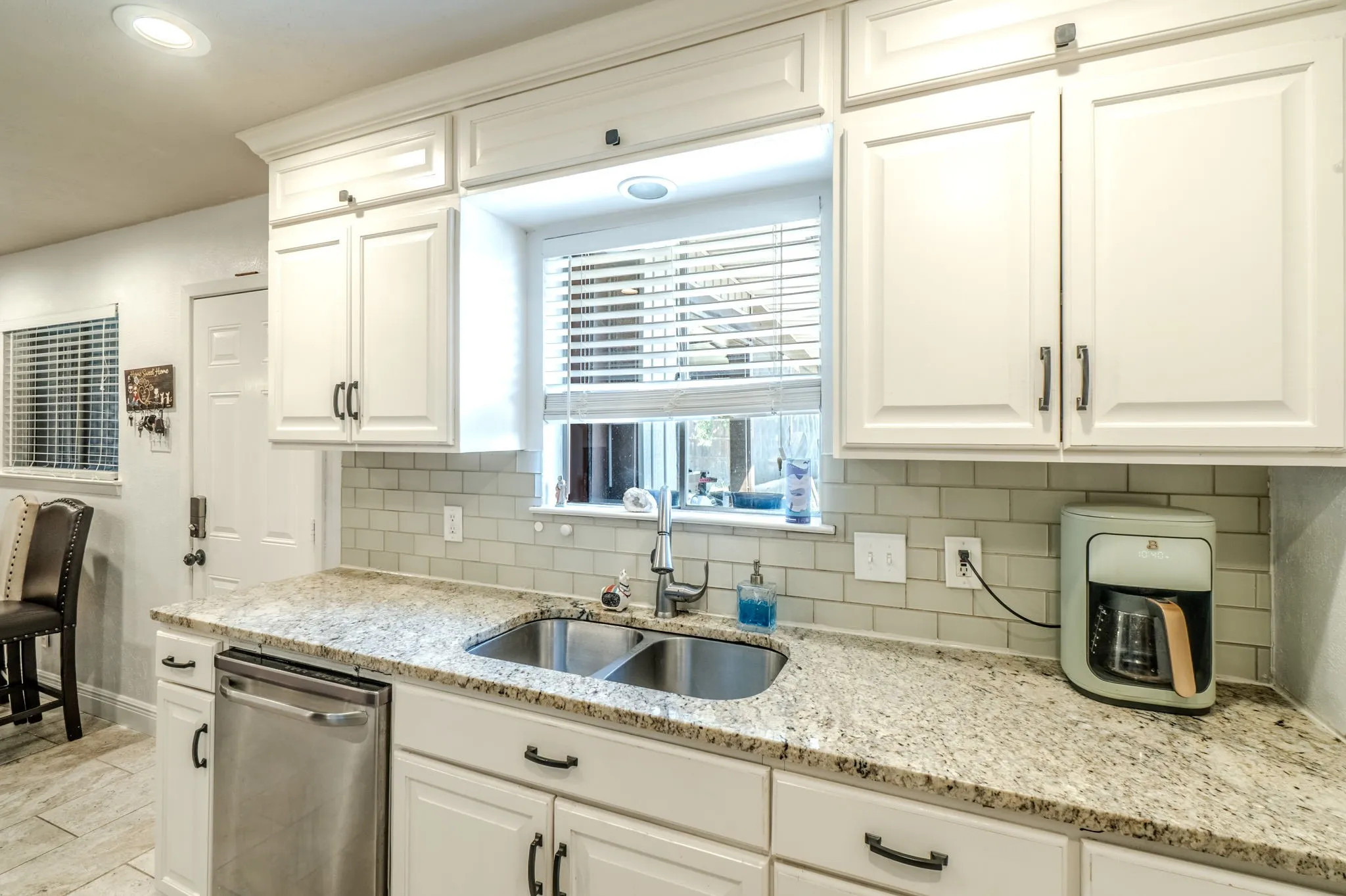 White shaker-style cabinetry with dark hardware, stainless steel sink, light-toned granite countertops, light subway tile backsplash, and a built-in stainless steel dishwasher