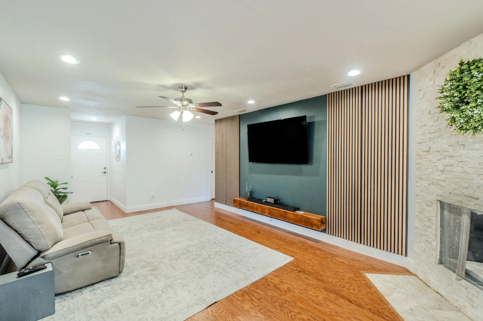 Wood-finish flooring, recessed lighting, ceiling fan, white paneled entry door with frosted glass insert, and a stone-clad fireplace