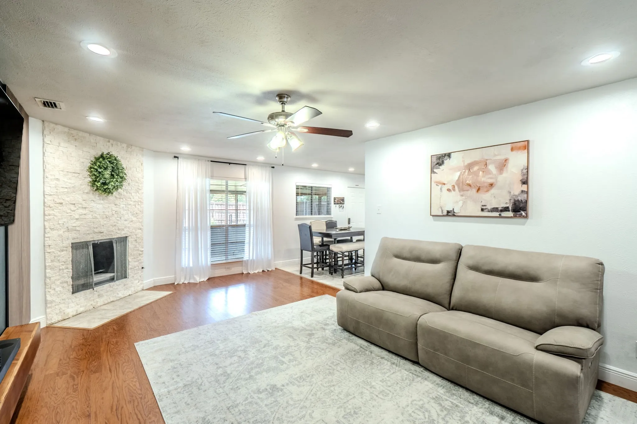 Hardwood-style flooring throughout, complemented by a stone-faced fireplace with a hearth, recessed ceiling lighting, and a ceiling fan