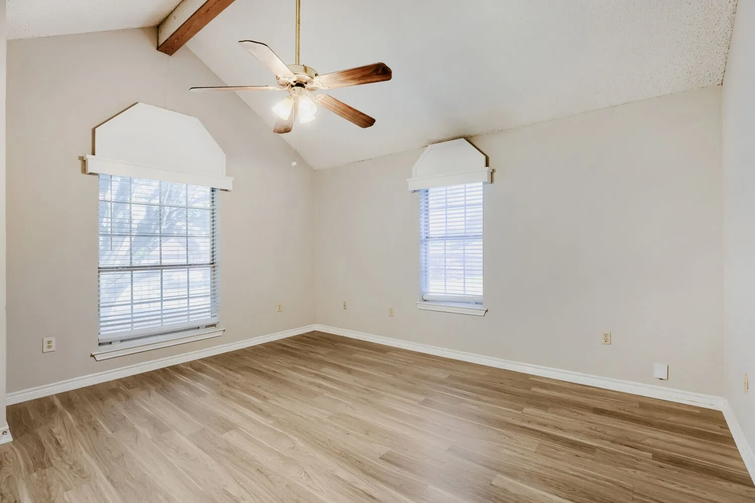 Vaulted ceiling with exposed wood beams, wood-finish flooring, two large windows with blinds, and an overhead ceiling fan