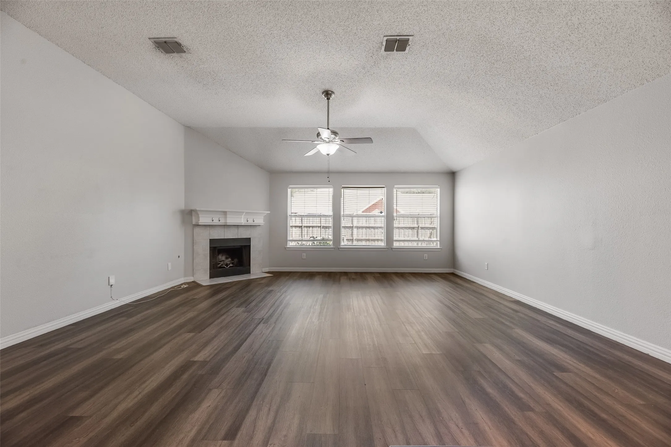 Spacious interior featuring vaulted ceilings, wood-finish flooring, a tile-surround fireplace with a mantel, a ceiling fan, and multiple windows