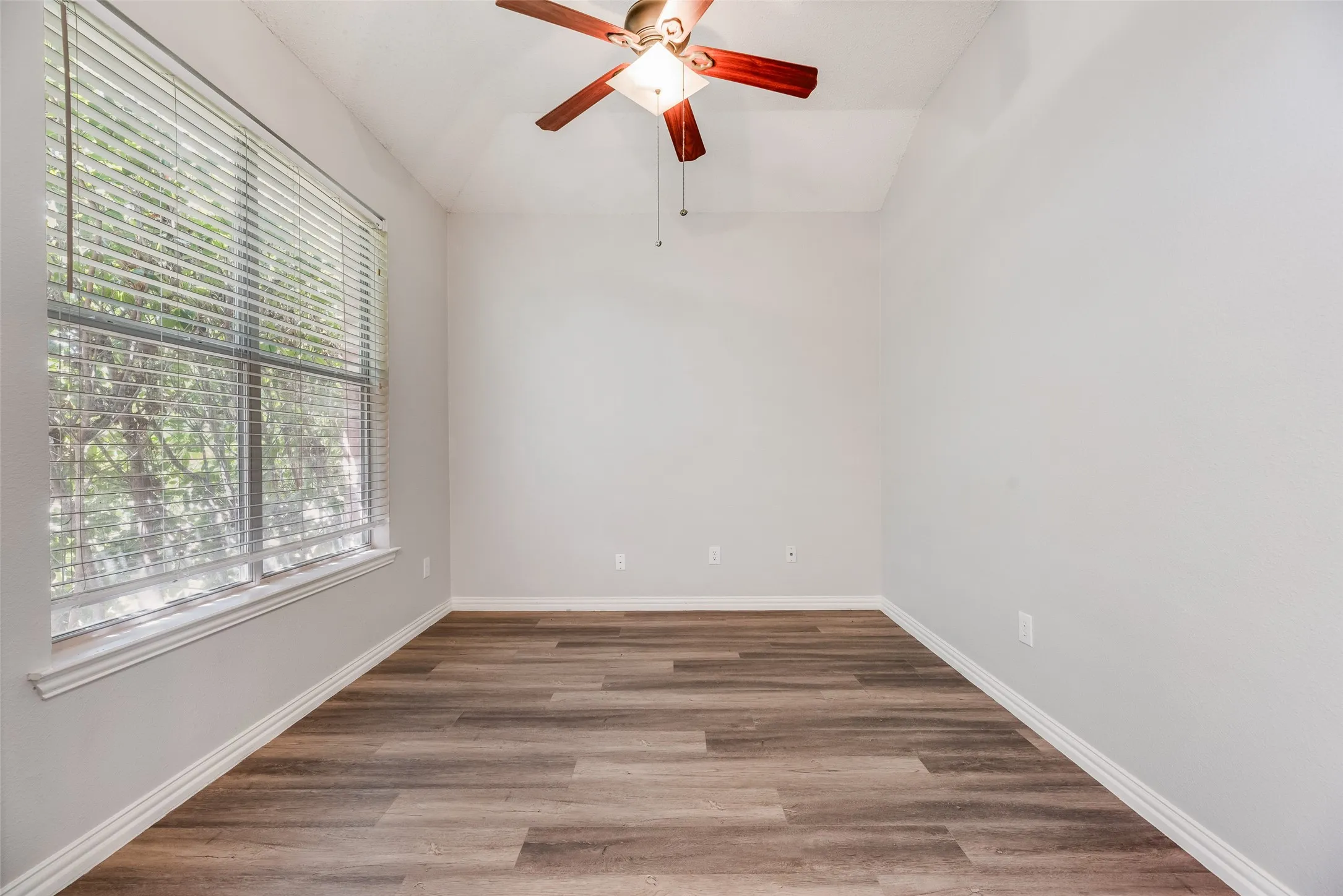 Vaulted ceiling with a ceiling fan, large window with blinds, wood-finish flooring, and white walls