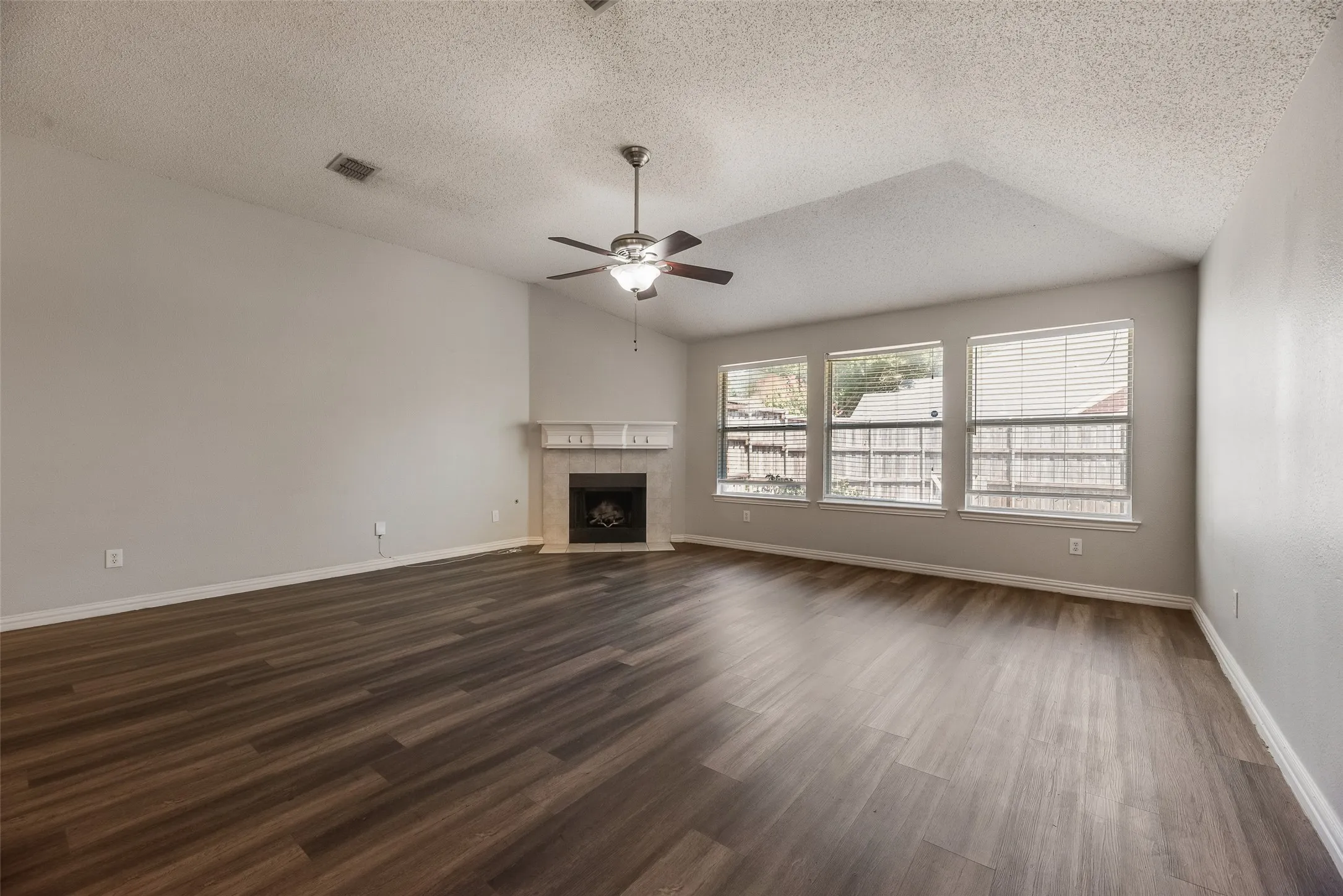 Spacious living area featuring wood-finish flooring, a vaulted ceiling, a ceiling fan, a fireplace with a stone surround and mantel, and three windows
