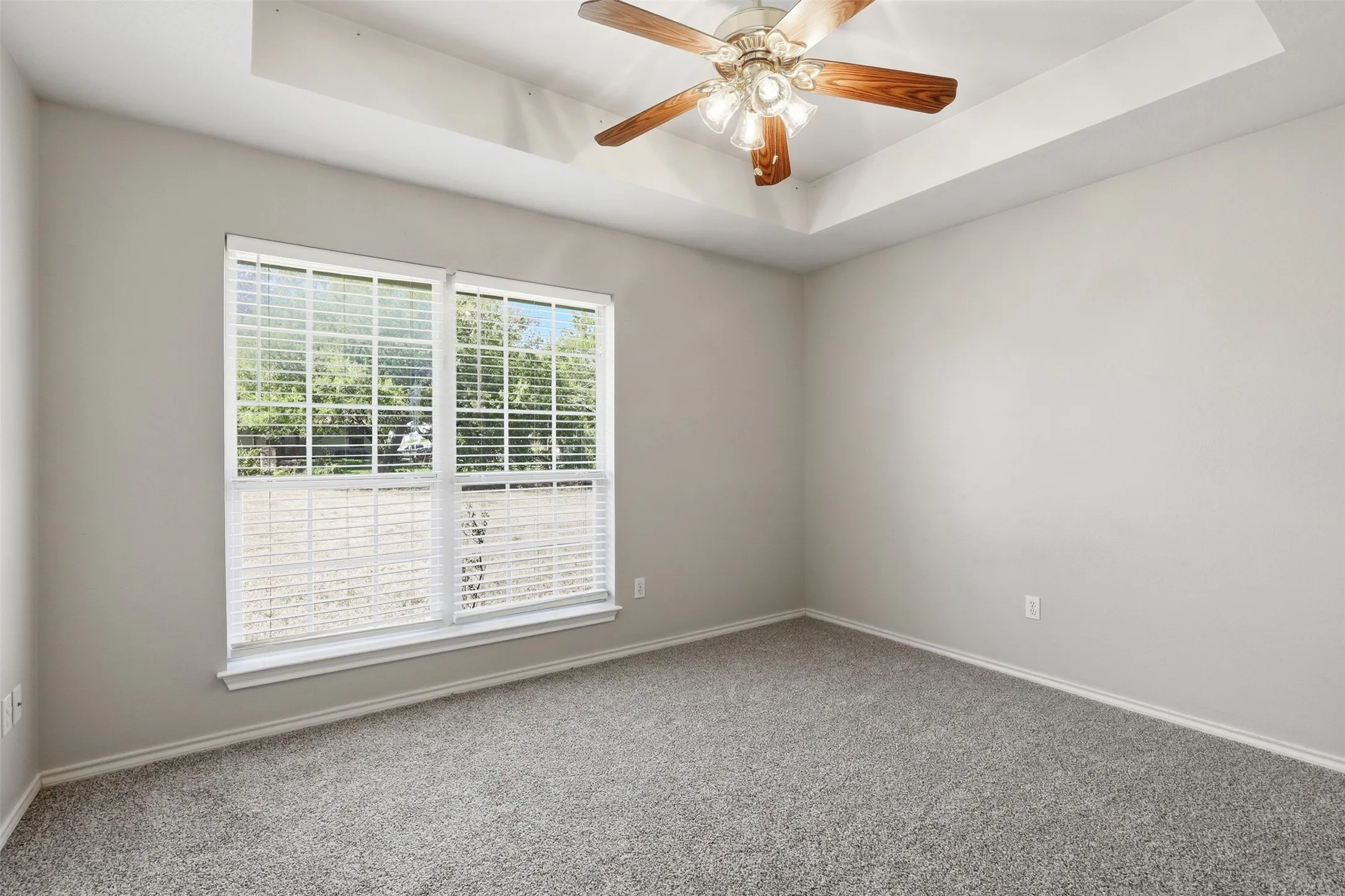 Bright bedroom showcasing neutral carpeting, a ceiling fan with wood-finish blades, and large dual-pane windows with plantation shutters providing natural light and privacy