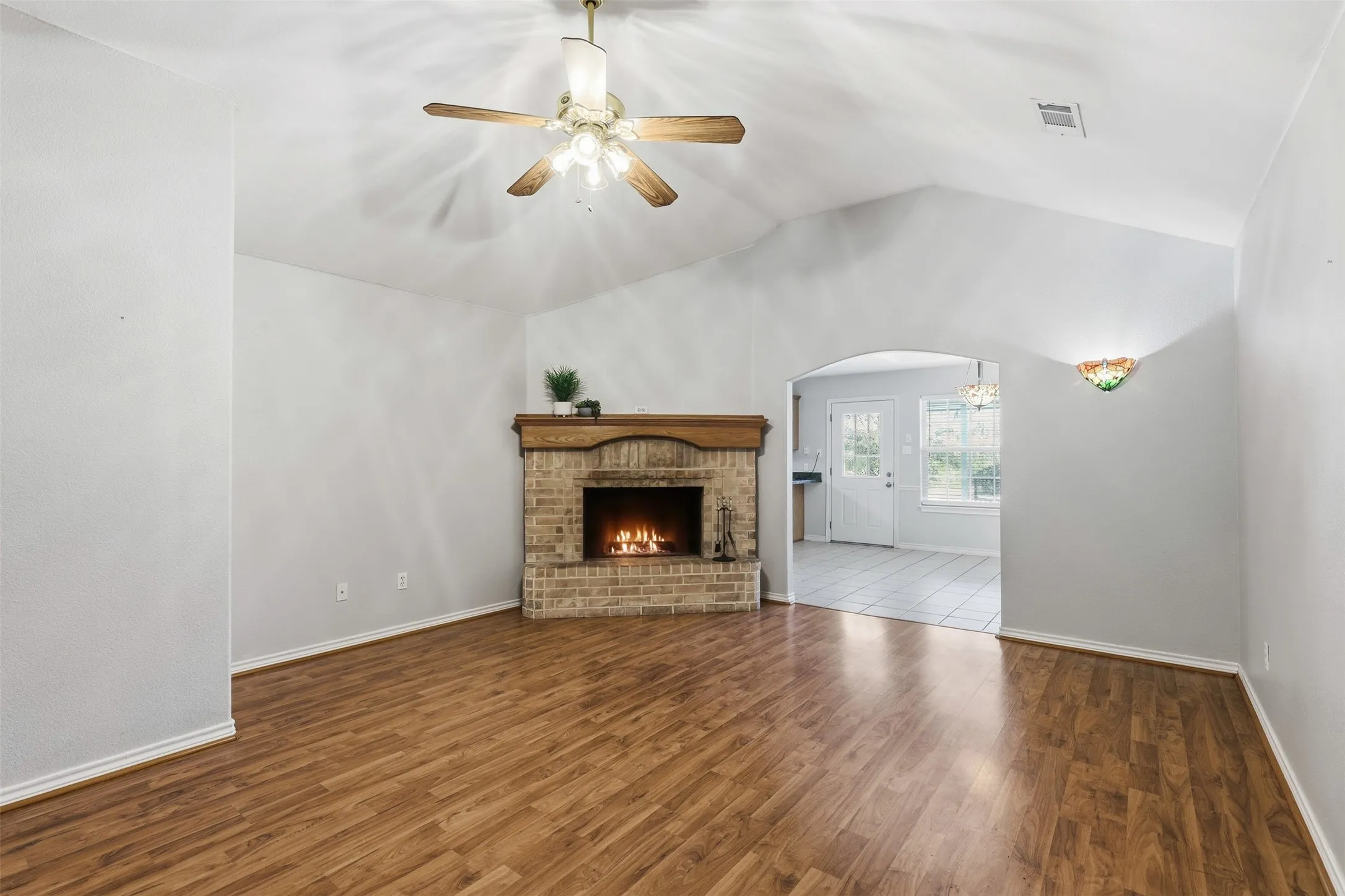 Vaulted ceiling with a ceiling fan, brick fireplace with a wood mantel, wood-finish flooring, and an arched doorway leading to a tiled area