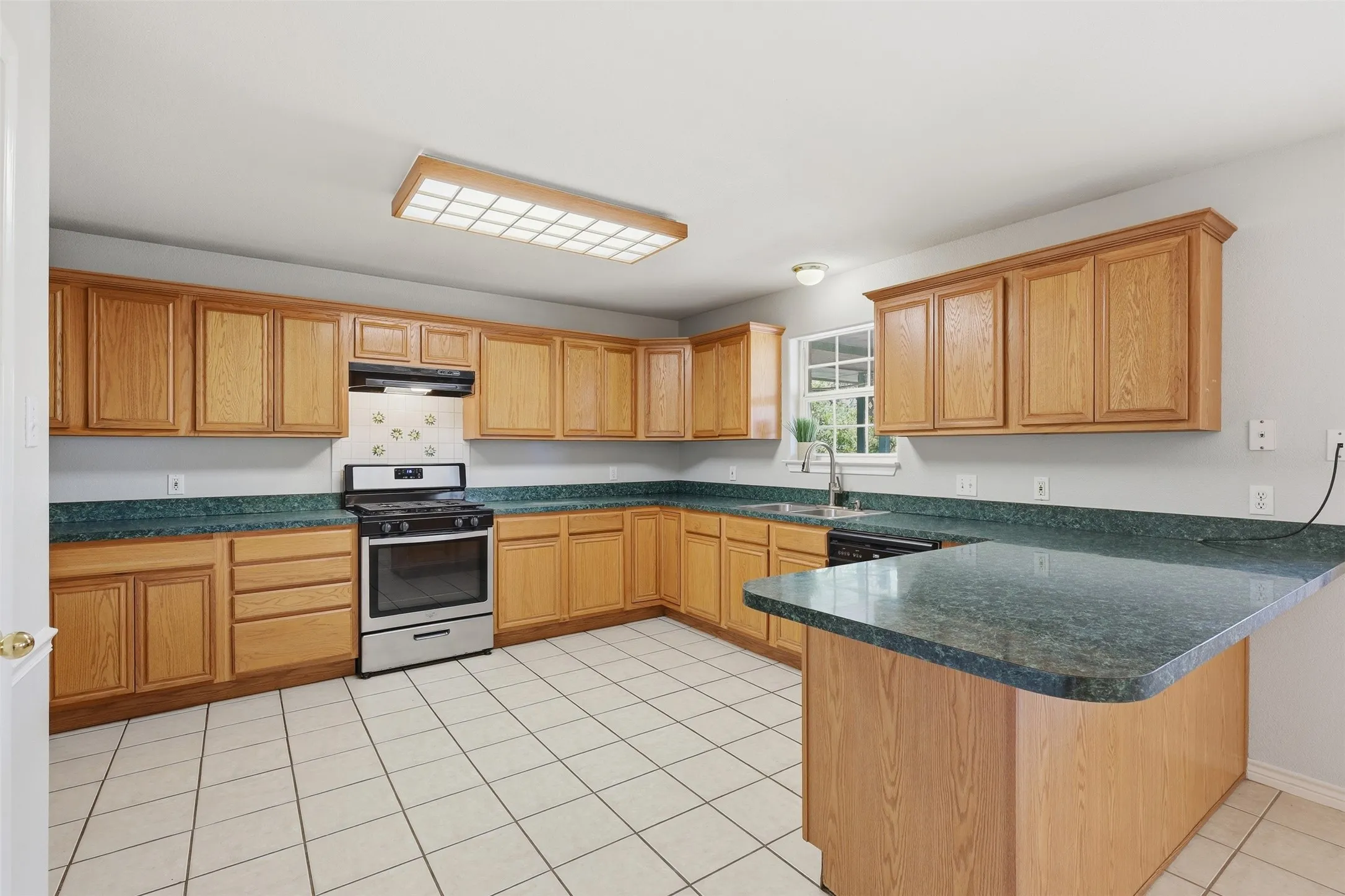 Spacious kitchen featuring warm wood cabinetry throughout, stainless steel appliances, green stone countertops, a large center island with wood-finish base, tile flooring, and abundant natural light from multiple windows