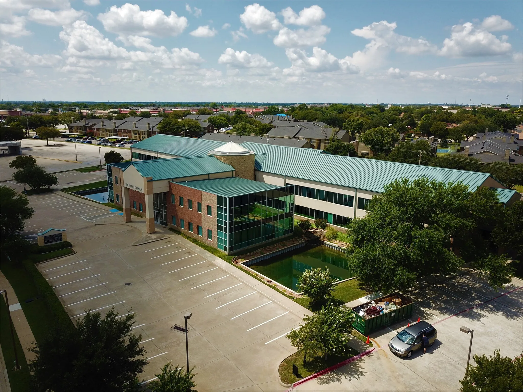 Building exterior featuring a multi-story glass facade, light brick accents, a prominent circular architectural element, and a teal-colored metal roof