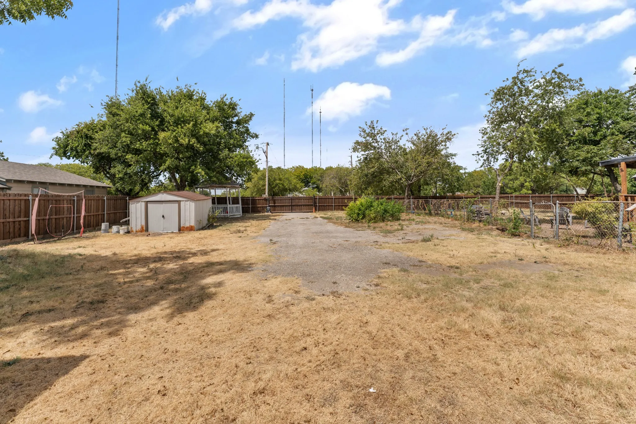 Expansive outdoor space featuring a storage shed, a gazebo, mature trees, and a gravel driveway