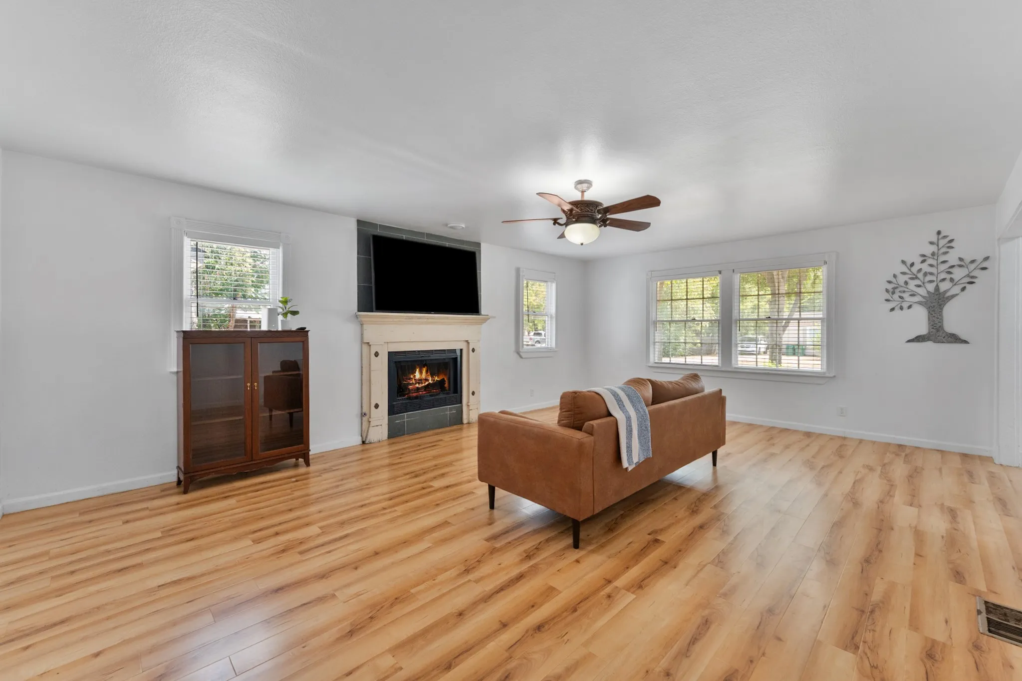 Light wood-finish flooring, a fireplace with a stone mantel, multiple windows, and a ceiling fan with integrated lighting