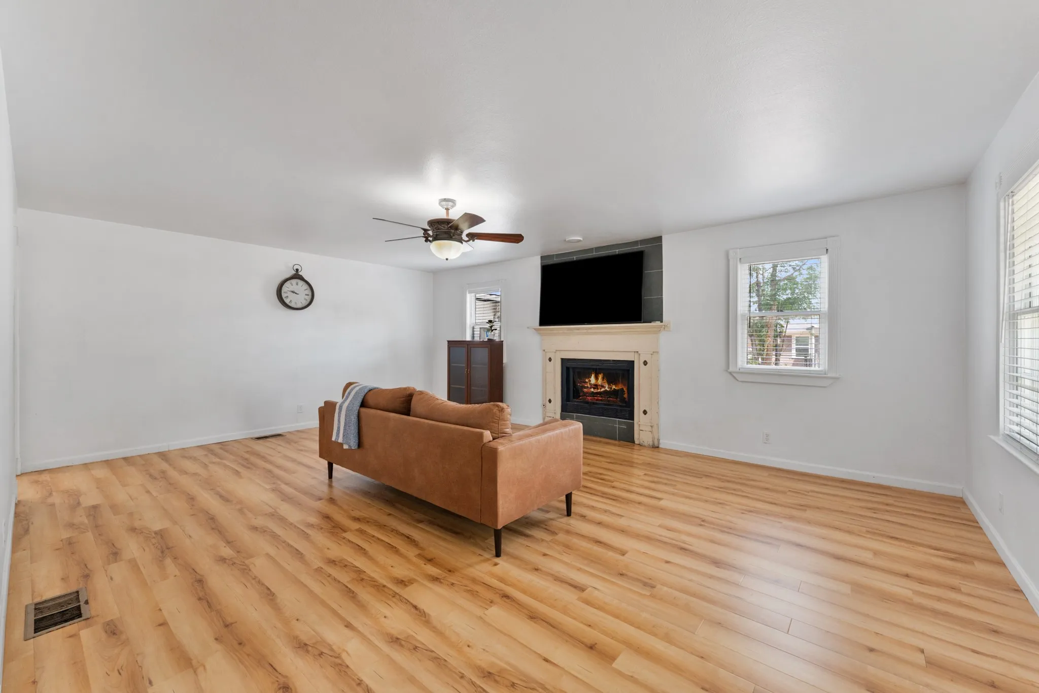 Spacious living area featuring wood-finish flooring, a ceiling fan with integrated lighting, and a fireplace with a stone surround