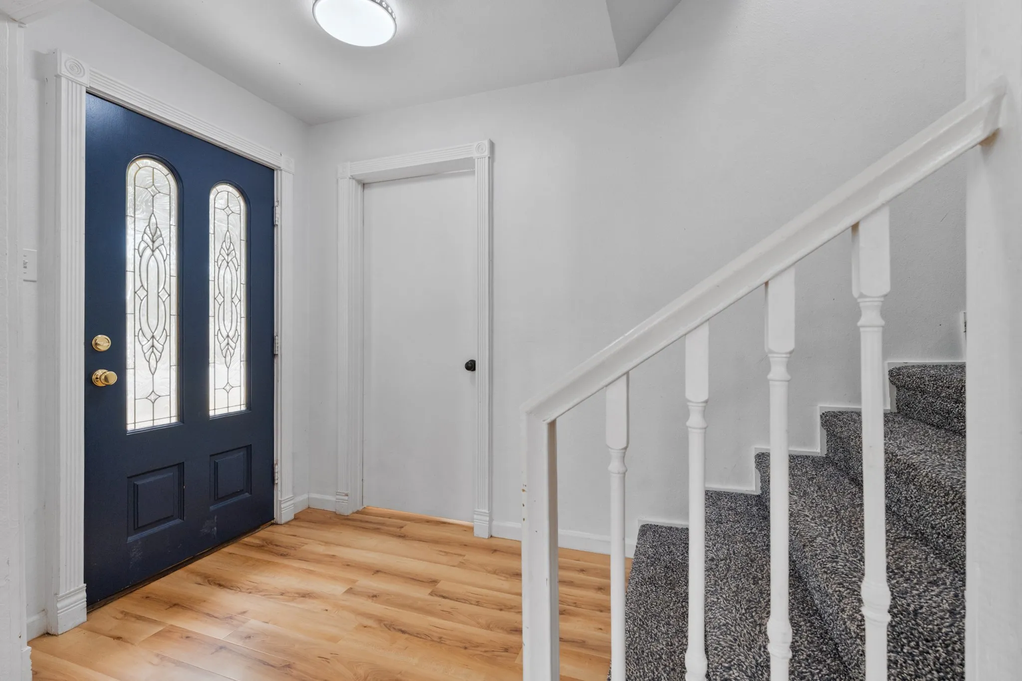 Entryway featuring a dark blue door with decorative glass inserts, wood-finish flooring, and a carpeted staircase with white balusters