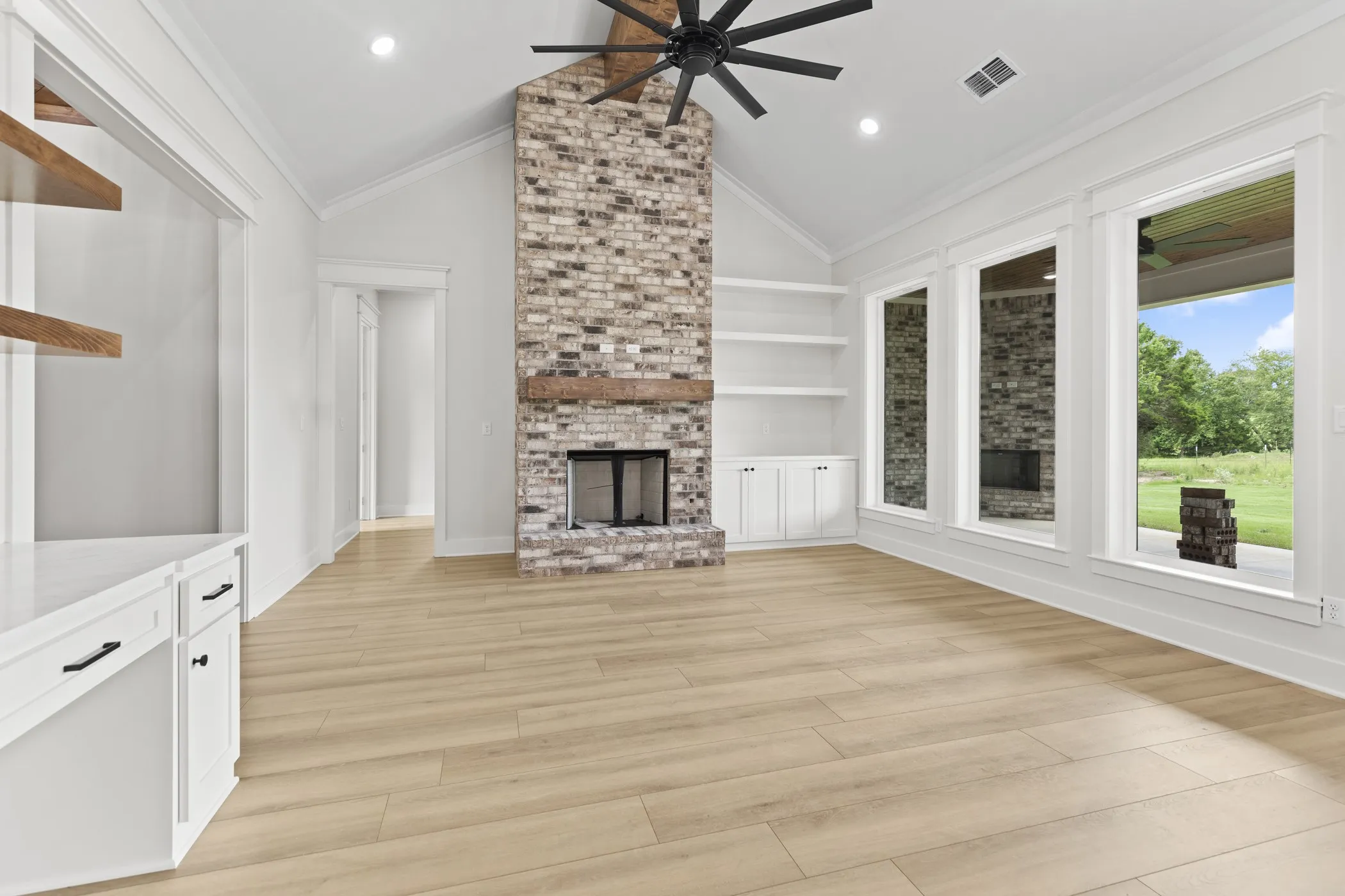 Vaulted ceiling with exposed beam fan, brick fireplace with wood mantel, built-in shelving and cabinetry, wood-finish flooring, and multiple large windows