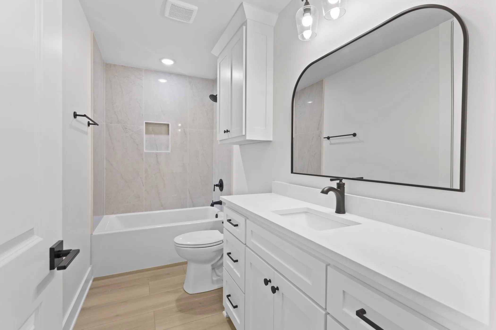 White vanity with integrated sink and matte black faucet, arched framed mirror, white-tiled shower with built-in niche, and wood-finish flooring
