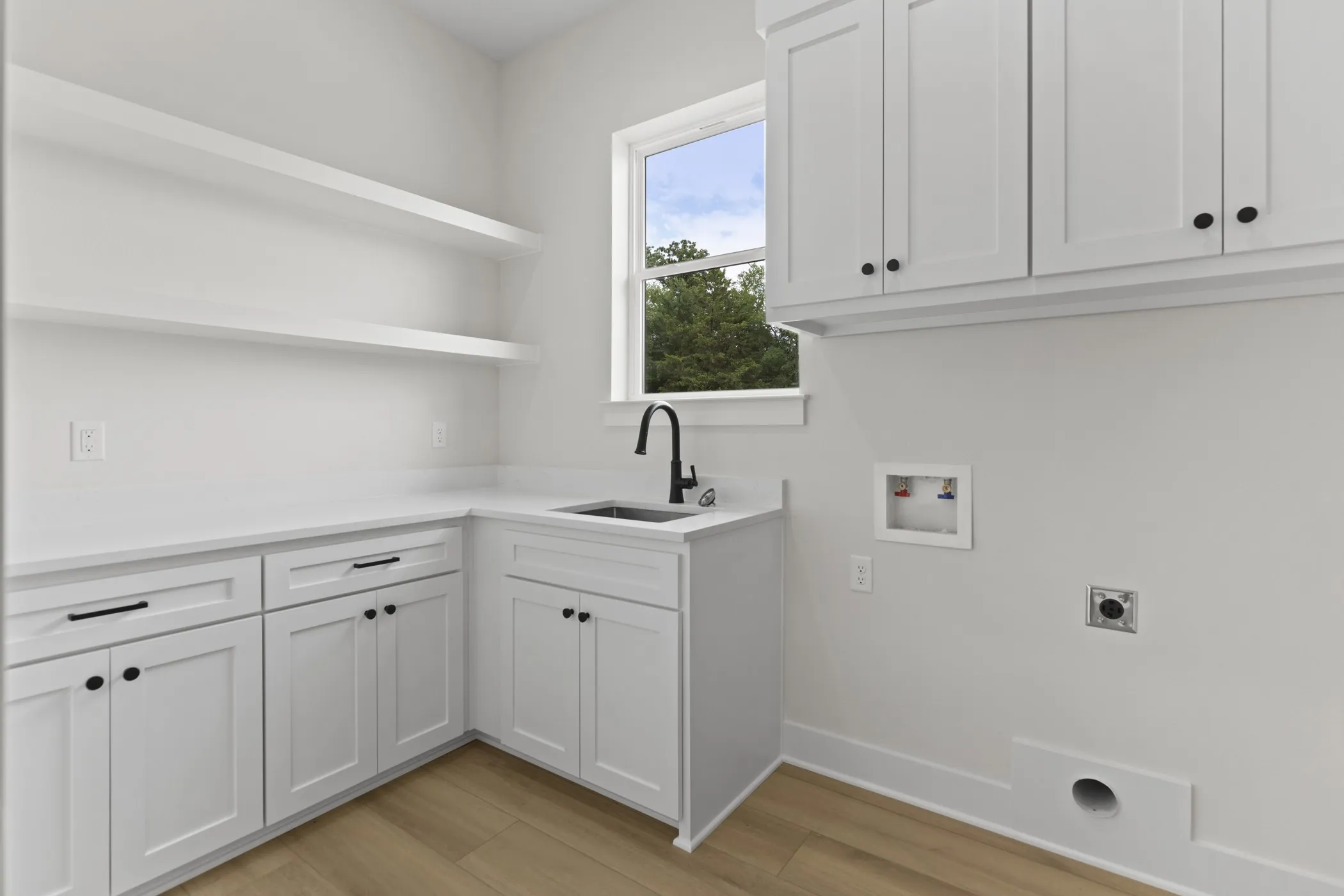 White shaker-style cabinetry, floating open shelving, light-toned wood-finish flooring, undermount sink with a matte black faucet, and white countertops