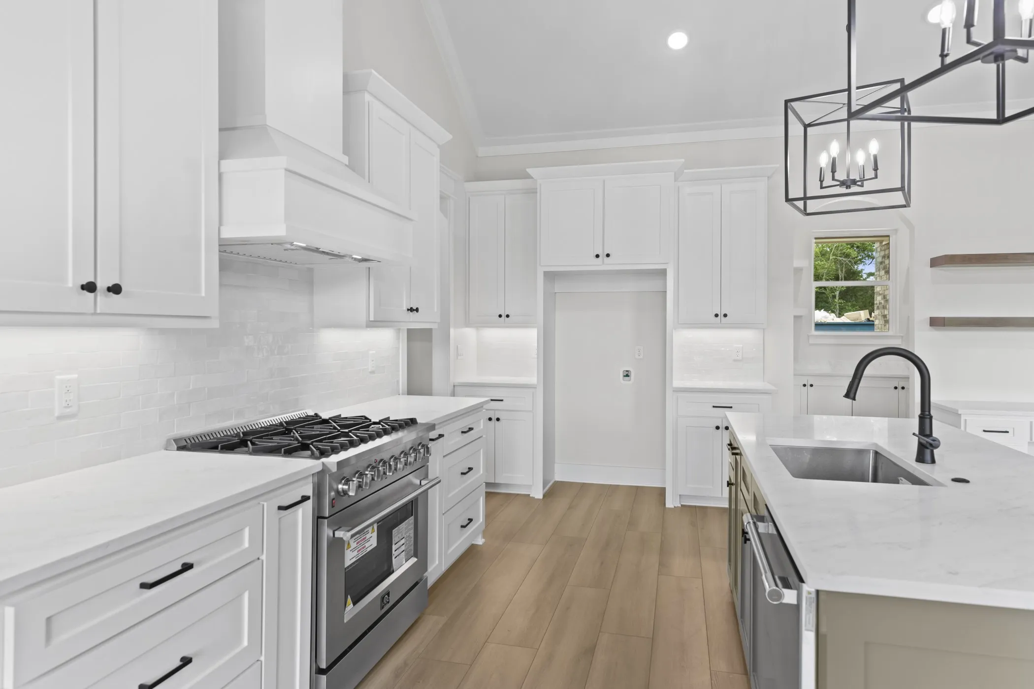 White shaker cabinetry, stainless steel range, light-toned wood-finish flooring, island with undermount sink, and contemporary chandelier