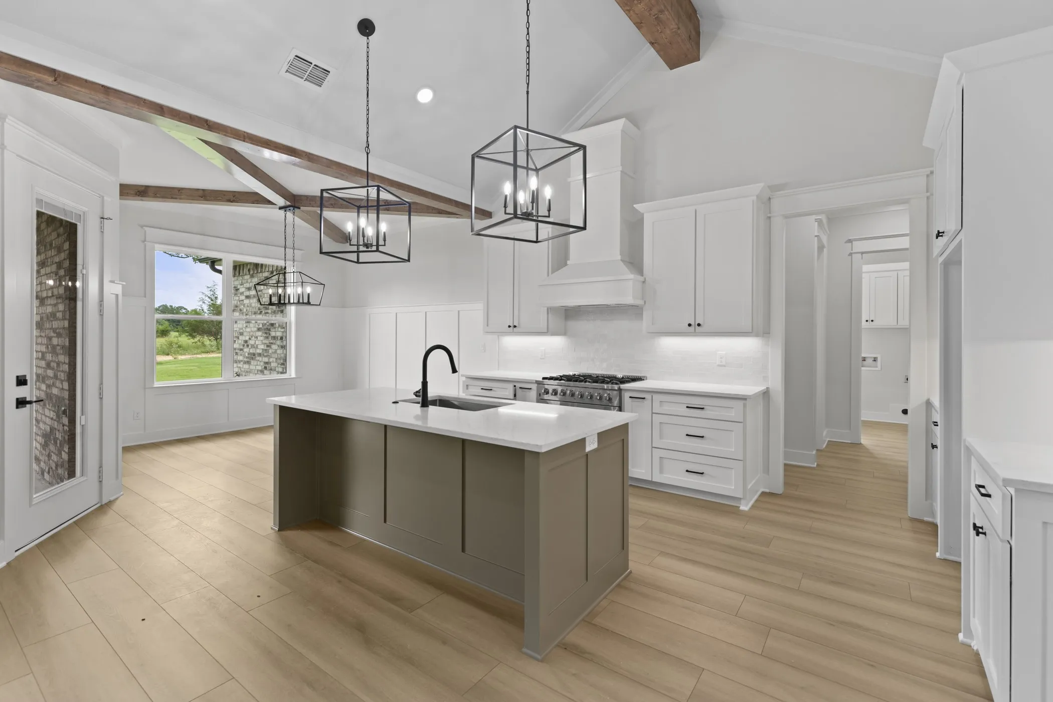 Vaulted ceiling with exposed wood beams, wood-finish flooring, central kitchen island with integrated sink, white shaker-style cabinetry, and stainless steel range with custom hood