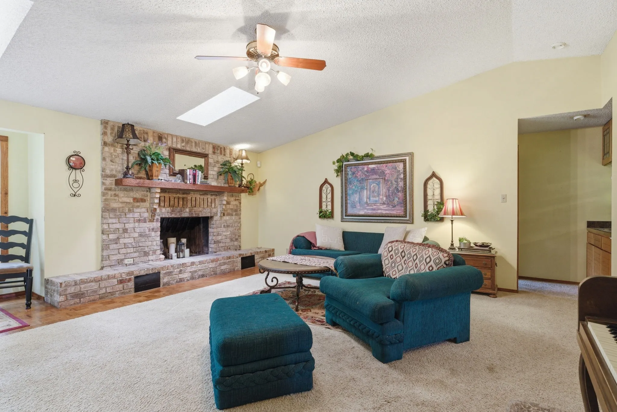Vaulted ceiling with skylight, brick fireplace with wood mantel, ceiling fan with light fixture, and wood-finish flooring