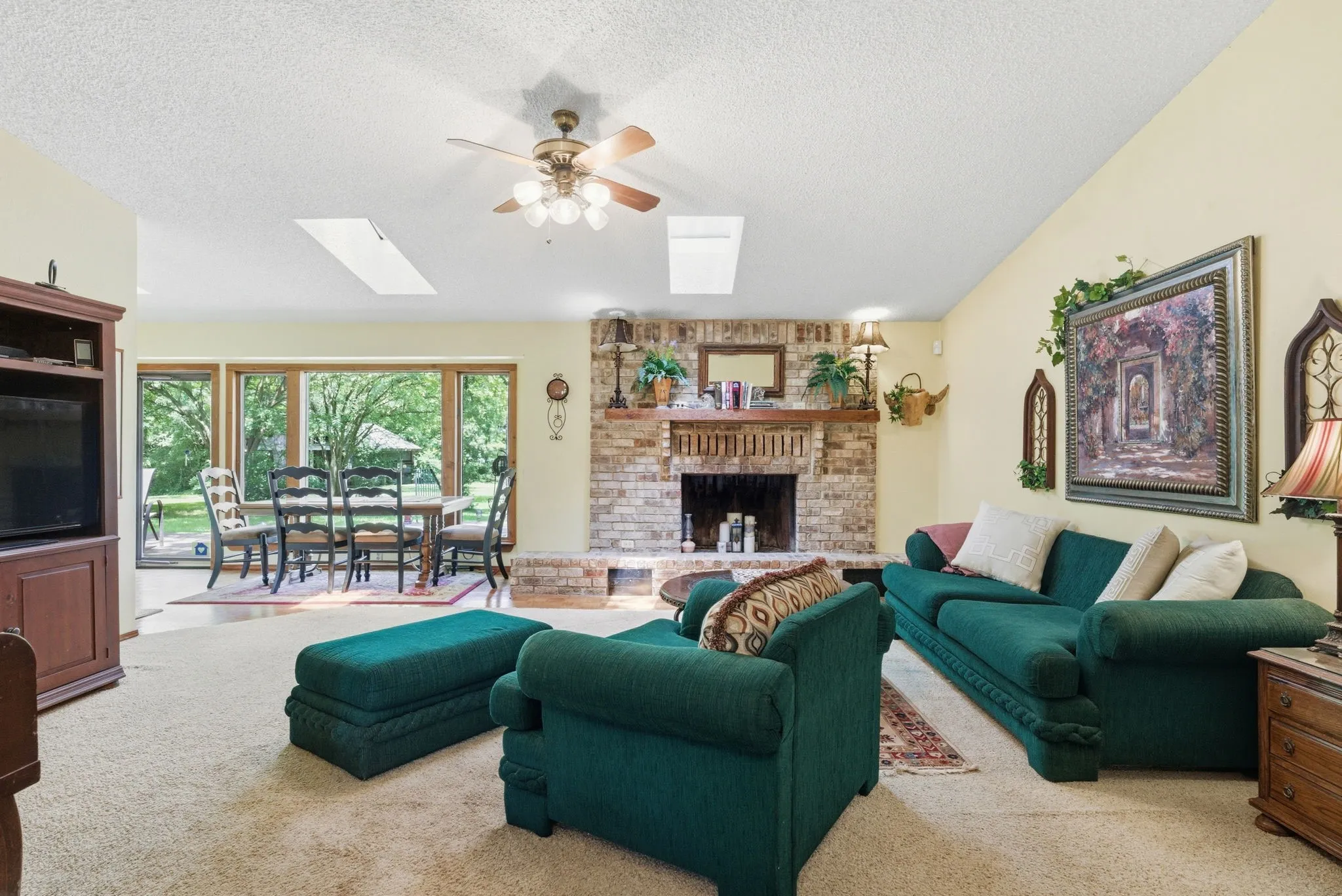 Brick fireplace with a wood-finish mantel, skylights, ceiling fan with integrated lighting, and expansive windows