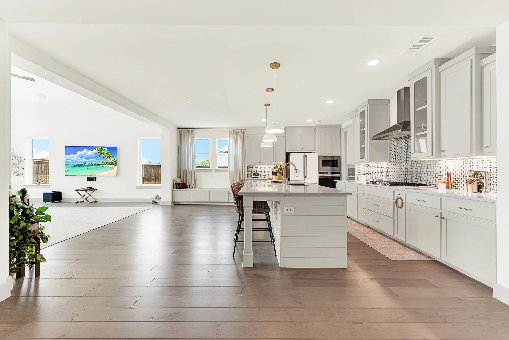 A long sightline spans across warm hardwood floors into a sunny wall of windows. It’s a welcoming spot where kids can finish homework at the counter while dinner simmers.