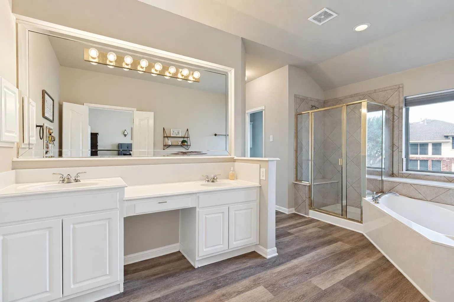 Dual vanity with integrated dressing table, oversized mirror with strip lighting, wood-finish flooring, tiled shower with glass enclosure, and a built-in soaking tub
