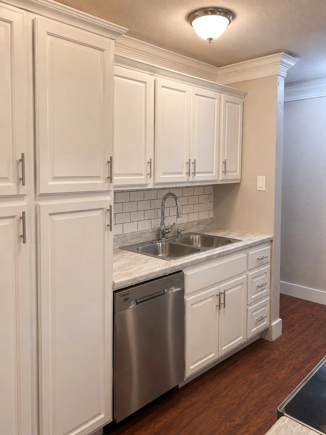White cabinetry with crown molding, stainless steel double basin sink, stainless steel dishwasher, white subway tile backsplash, and wood-finish flooring