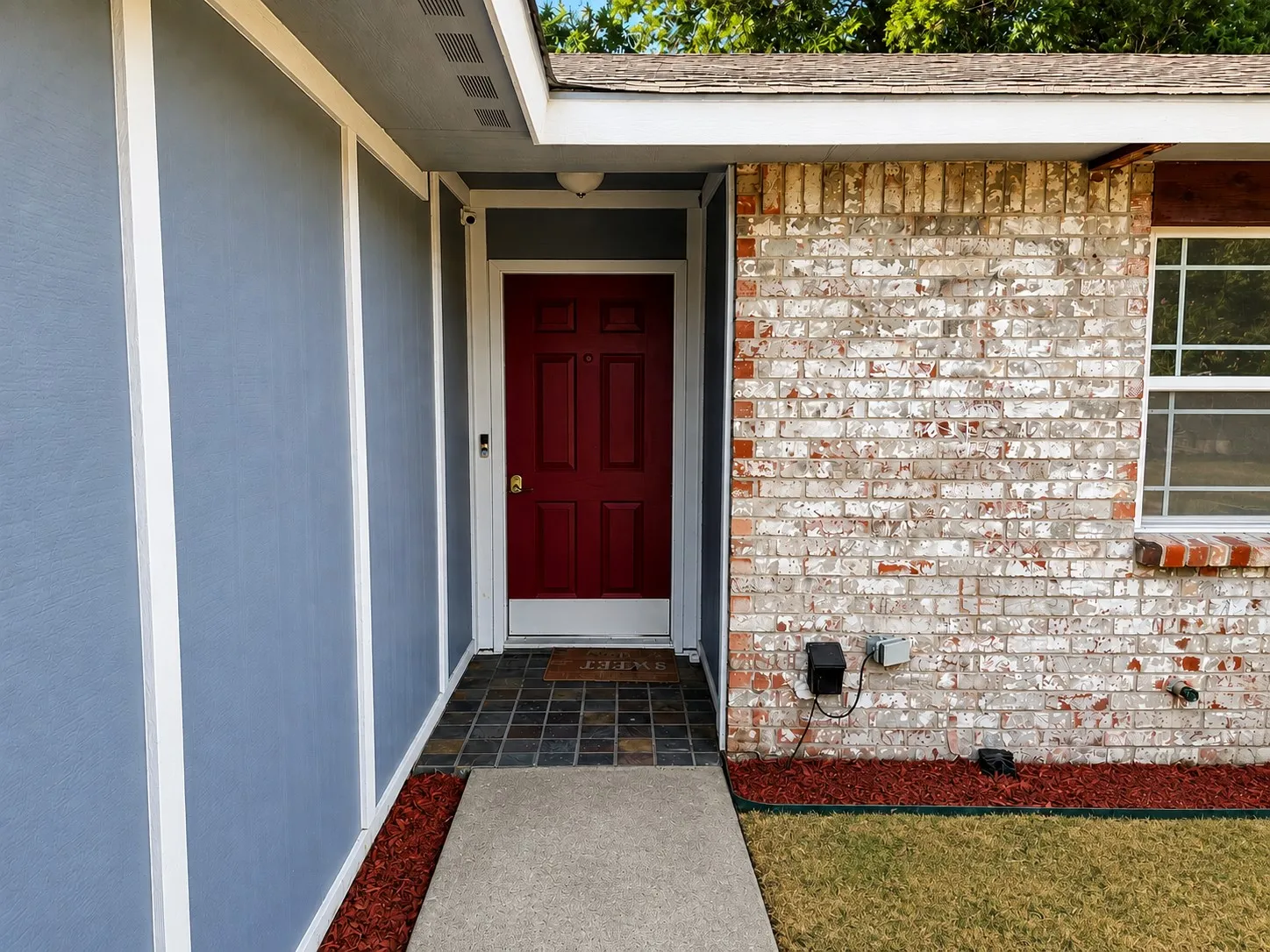 Brick exterior with distressed finish, red entry door, slate tile entry, concrete walkway, and landscaped red mulch