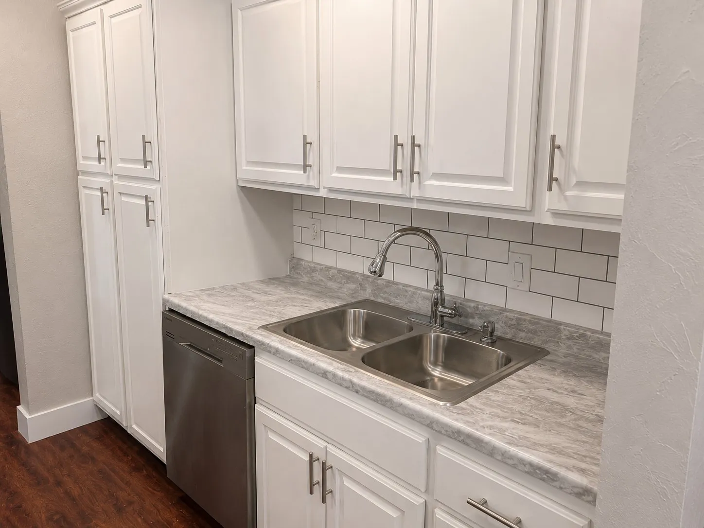 White shaker cabinetry, subway tile backsplash, dual basin stainless steel sink, stainless steel dishwasher, and wood-finish flooring