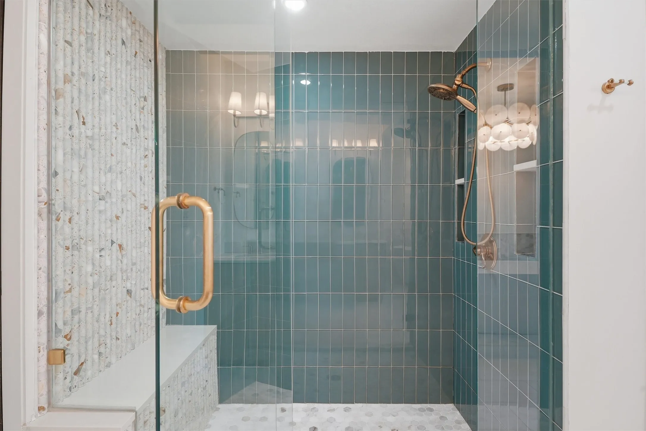 Frameless glass walk-in shower showcasing vertical teal subway tile, terrazzo accents, and a built-in bench. Outfitted with high-end brass hardware, a handheld shower sprayer, and a recessed storage niche.
