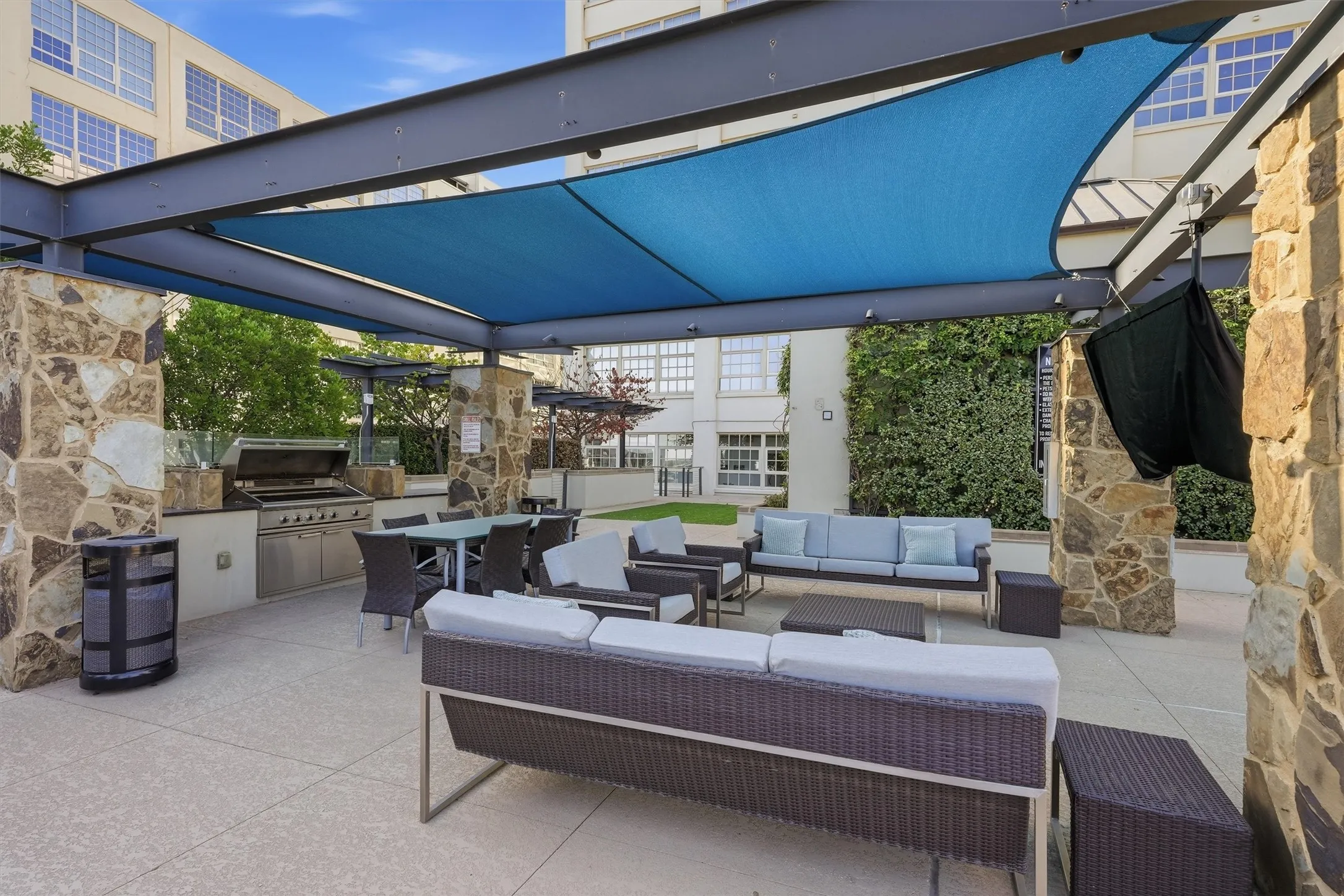 Covered outdoor pavilion on the amenity deck featuring a built-in stainless steel gas grill, outdoor dining table, and deep-seated lounge sofas. Designed with shade sails, stone pillars, and an outdoor TV for year-round outdoor entertaining.