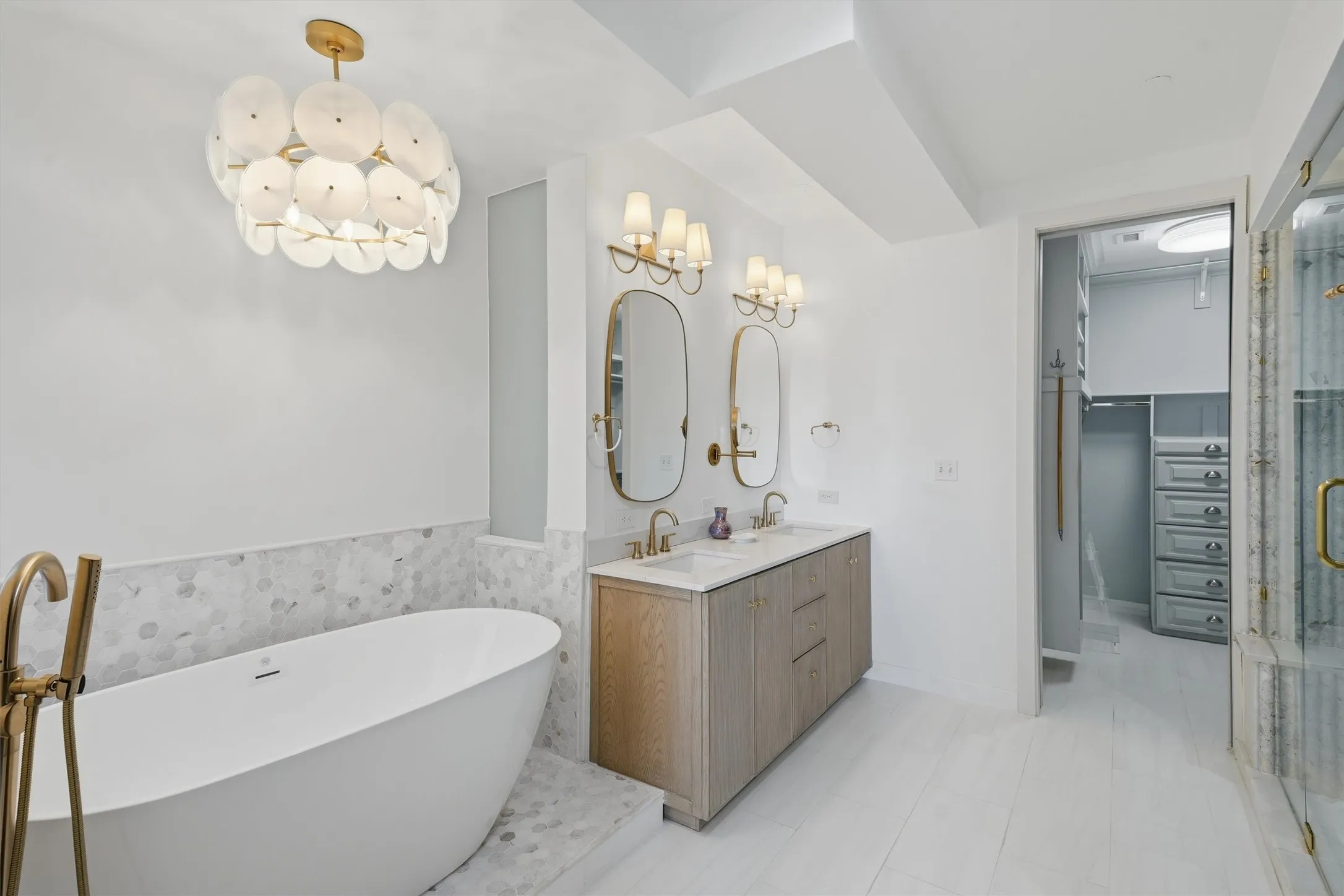 Spa-inspired primary bathroom featuring a freestanding soaking tub with floor-mounted brass hardware, a custom light-wood dual vanity, and marble hexagon tile accents. Connects directly to a spacious walk-in closet with built-in storage.