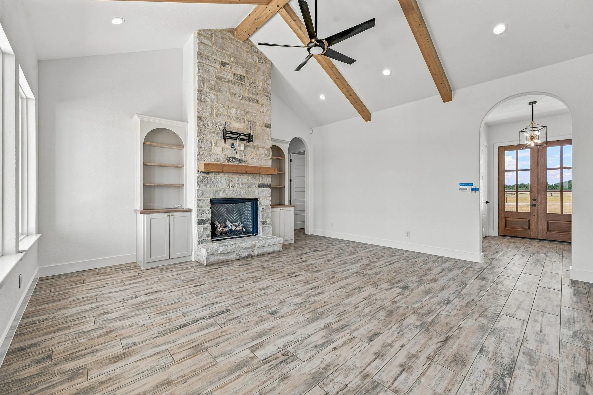 Vaulted ceiling with exposed wood beams, stone-clad fireplace, integrated built-in shelving, wood-finish flooring, and arched doorways