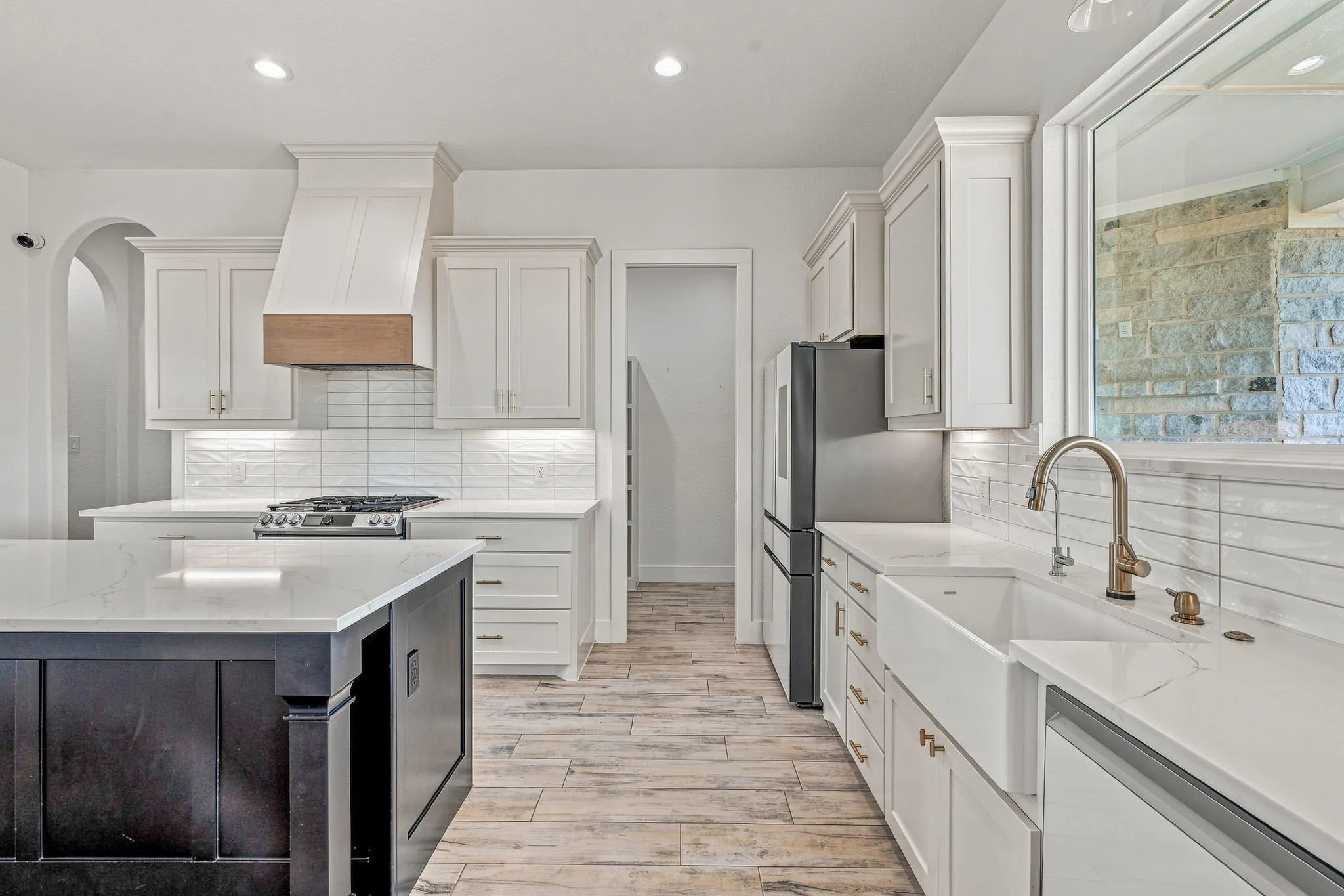 Kitchen featuring a large island with a light-toned countertop, white cabinetry with gold-toned hardware, a farmhouse-style sink with a brass-finish faucet, and wood-finish flooring