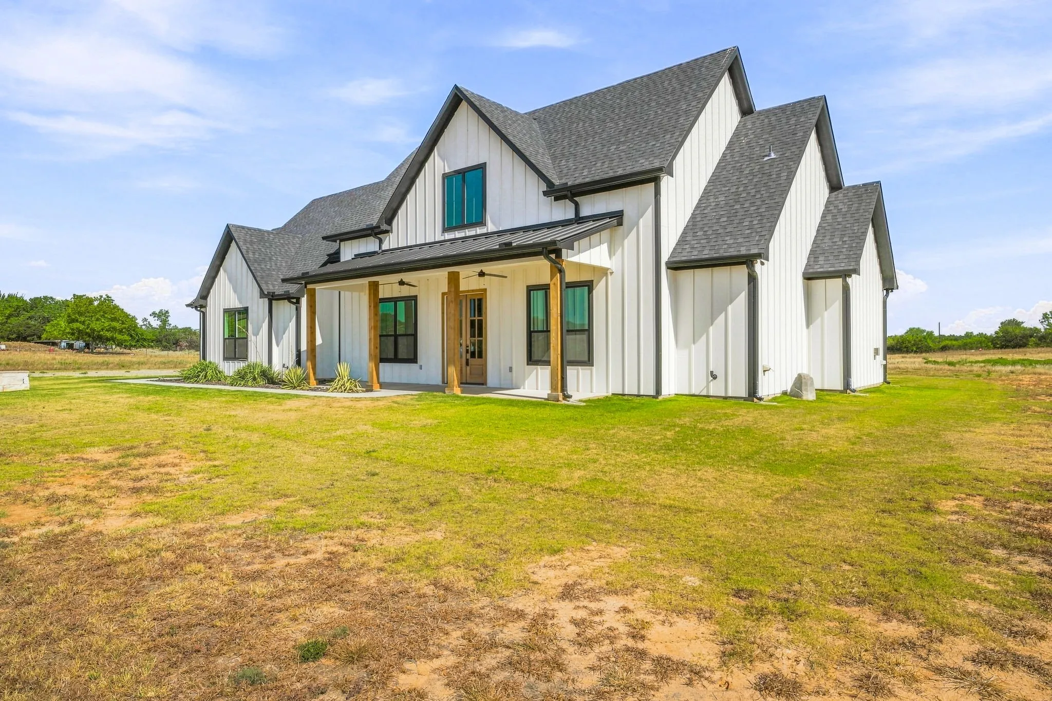 Modern farmhouse architecture with vertical white siding, a dark shingle roof, a covered porch with wood-tone columns, and dark-framed windows