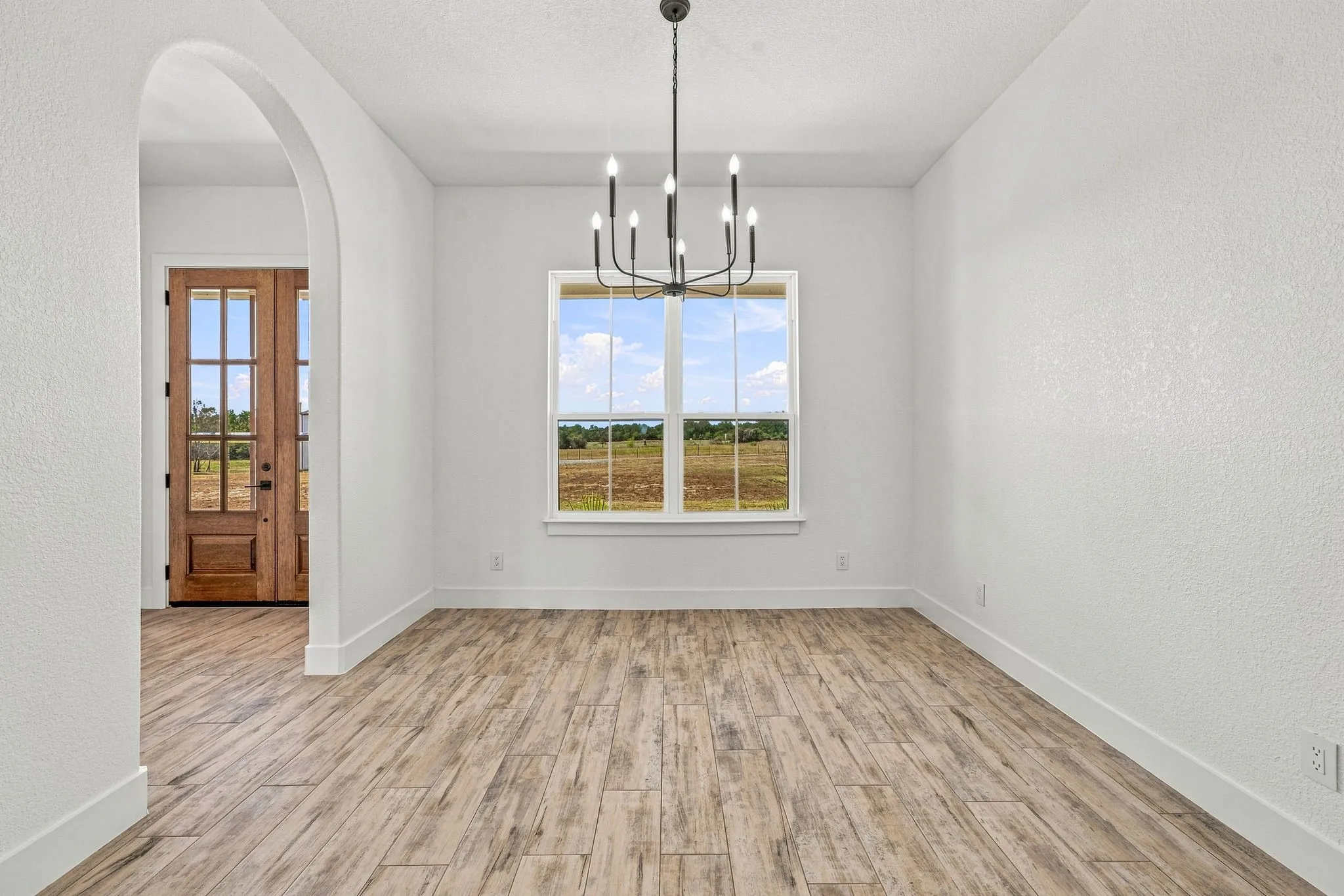 Arched entryway leading to a wood-paneled door with glass inserts, wood-finish flooring, a contemporary chandelier, and a large window
