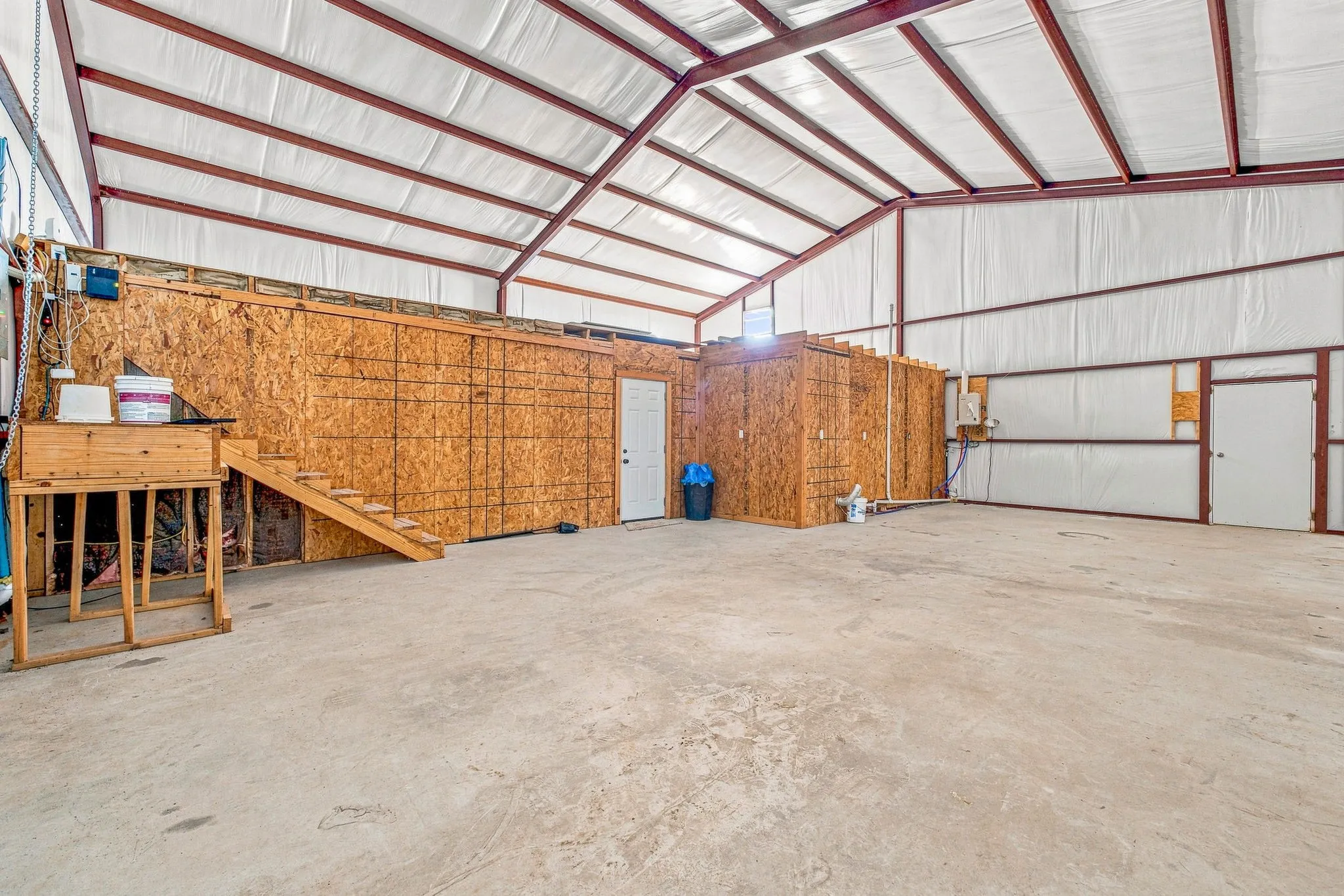Spacious interior featuring a concrete floor, exposed metal beam ceiling, and built-in plywood walls with a doorway and stairway access