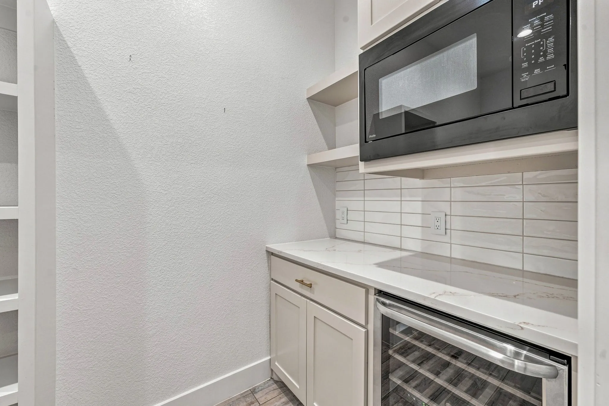 Built-in microwave oven, under-counter beverage refrigerator, light-toned cabinetry with brass hardware, white horizontal tile backsplash, and light-toned countertops