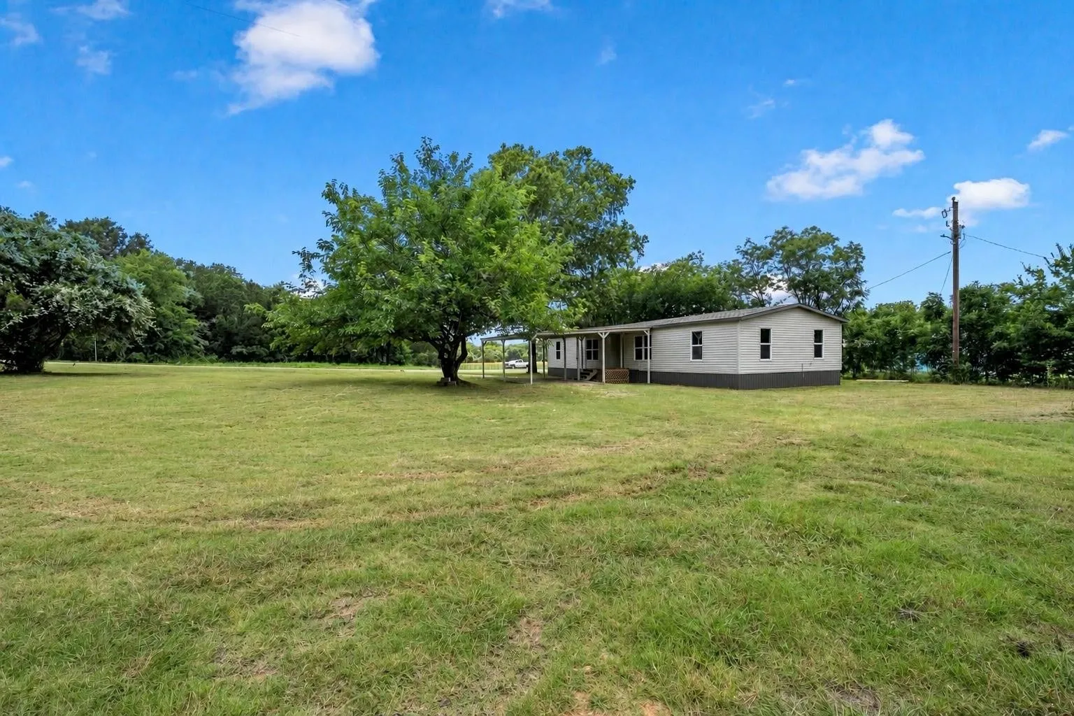 Expansive grassy property with scattered mature trees and a single-story residence featuring light siding and a covered porch