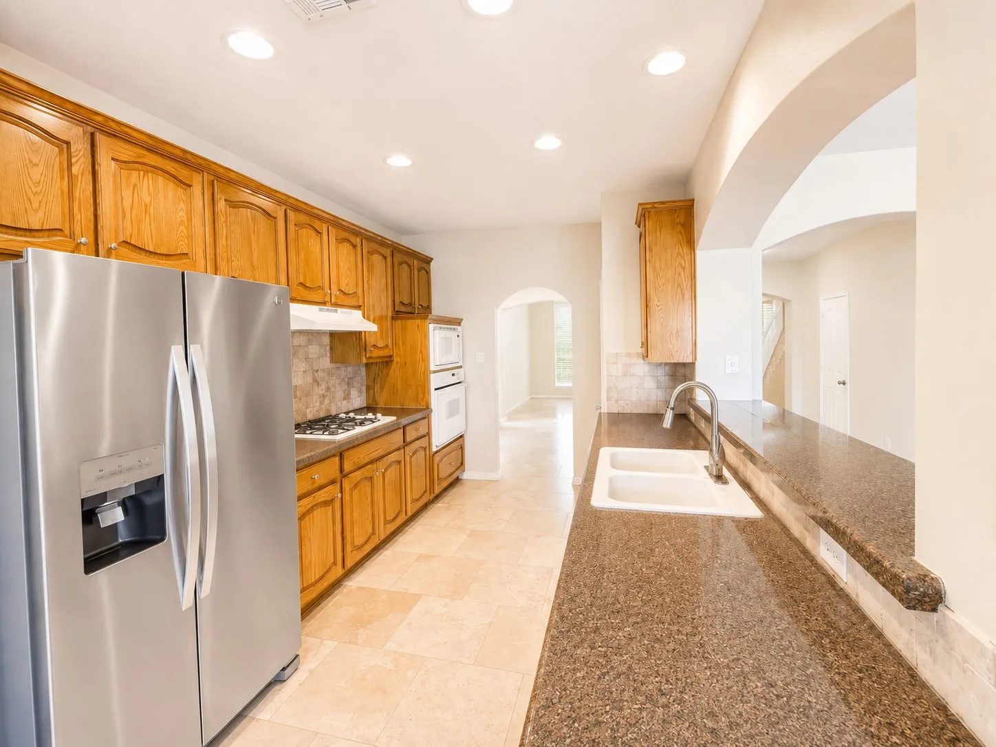 Wood-finish cabinetry, stainless steel refrigerator, integrated double oven, gas cooktop, and neutral tone tile flooring