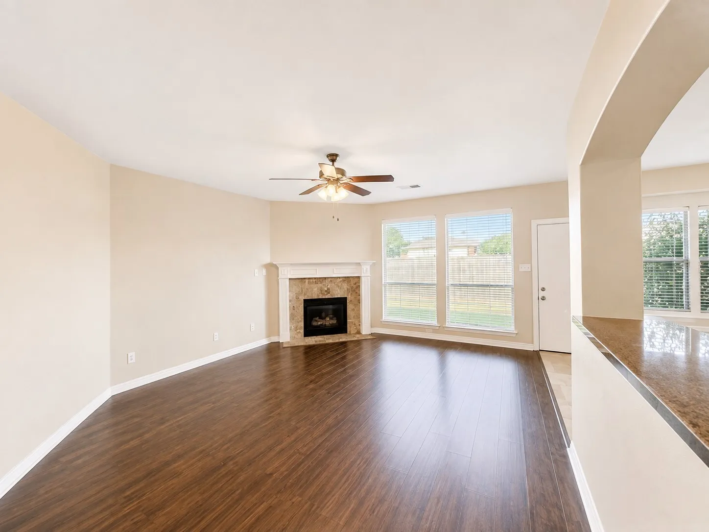 Spacious living area featuring rich wood-finish flooring, a corner fireplace with stone surround, a ceiling fan, and multiple windows