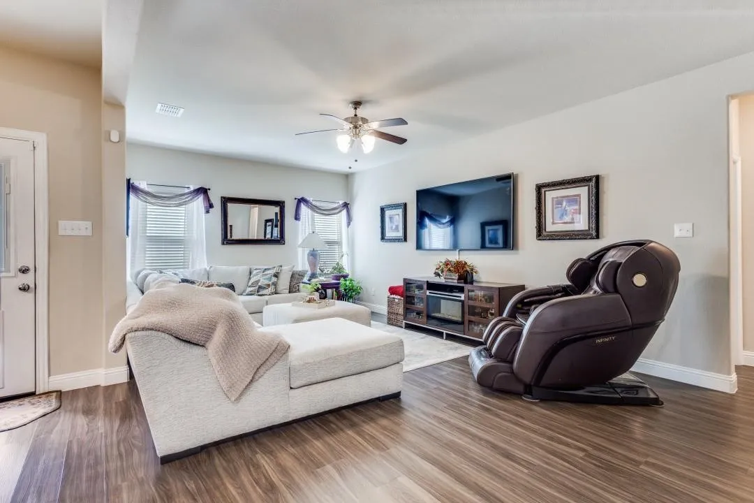 Spacious living area featuring wood-finish flooring, a ceiling fan, and multiple windows with blinds