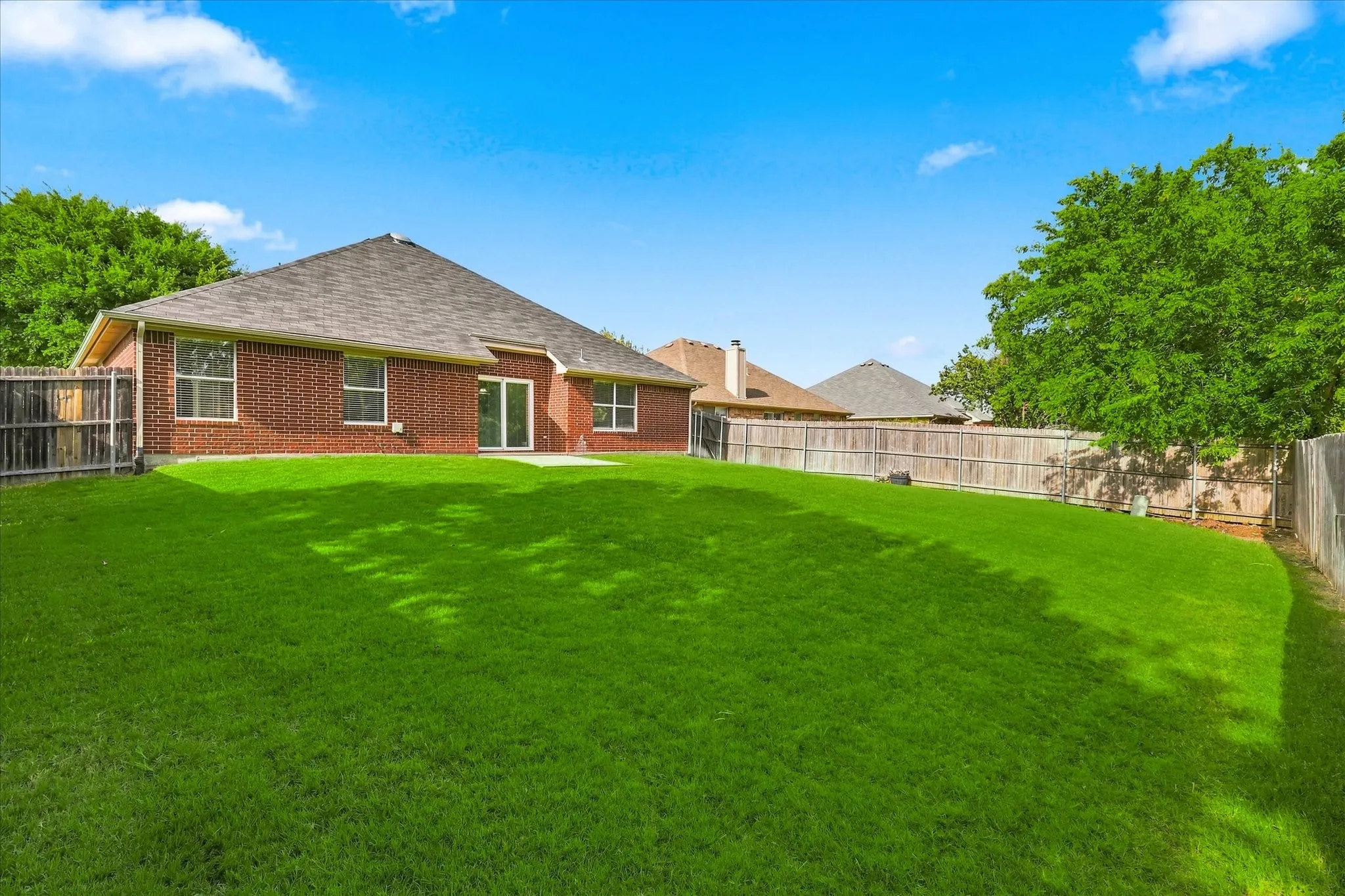 Expansive backyard featuring a manicured lawn, full privacy fencing, mature trees, a brick exterior, and a built-in patio