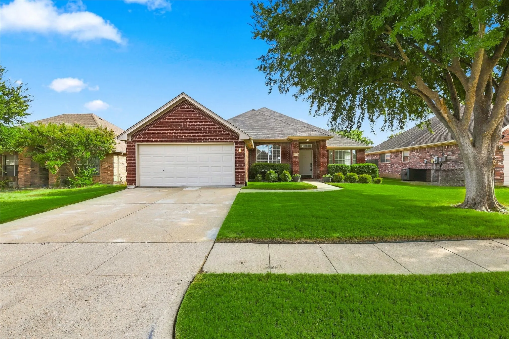 Red brick exterior, attached two-car garage, concrete driveway, mature tree, and landscaped front yard