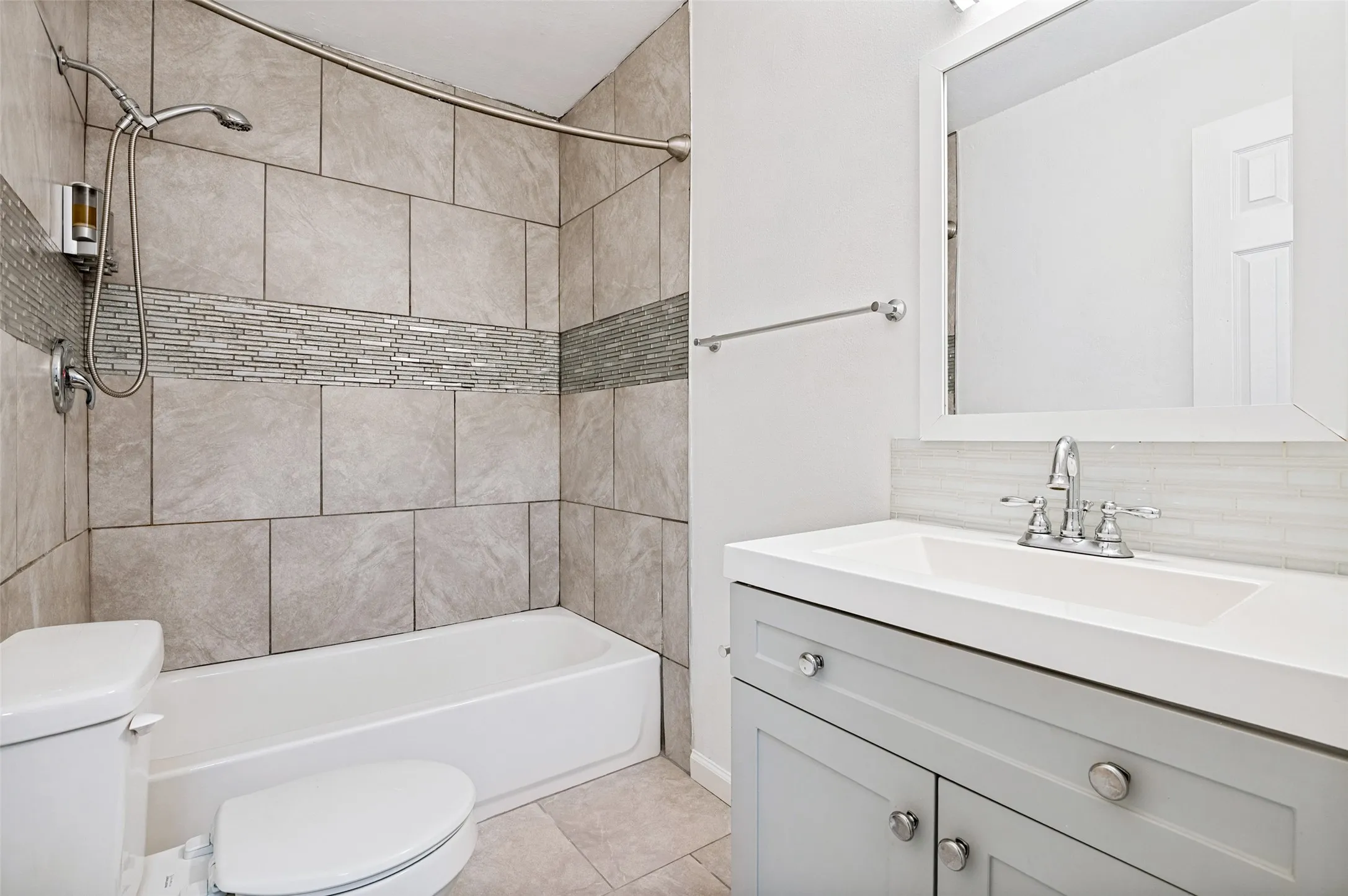 Bathroom featuring a vanity with a white countertop and undermount sink, a framed mirror, and a shower-over-tub with tile surround and decorative accent band