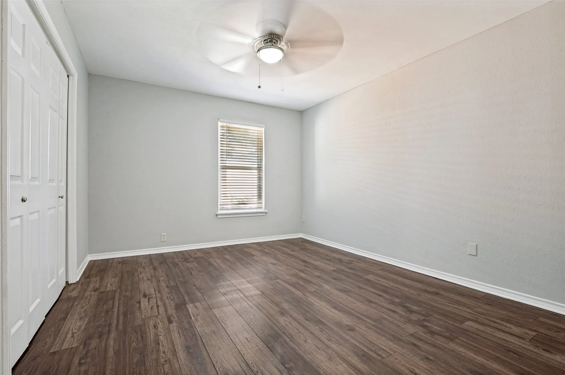 Wood-finish flooring, white bi-fold closet doors, window with blinds, and a ceiling fan with integrated lighting