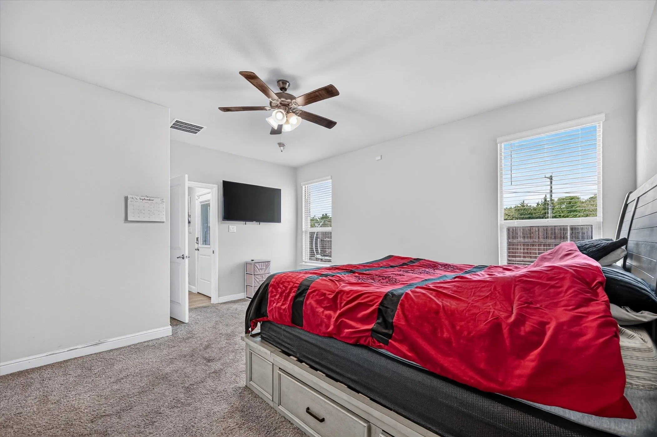 Carpeted room featuring a ceiling fan with light, two windows with blinds, and a mounted television