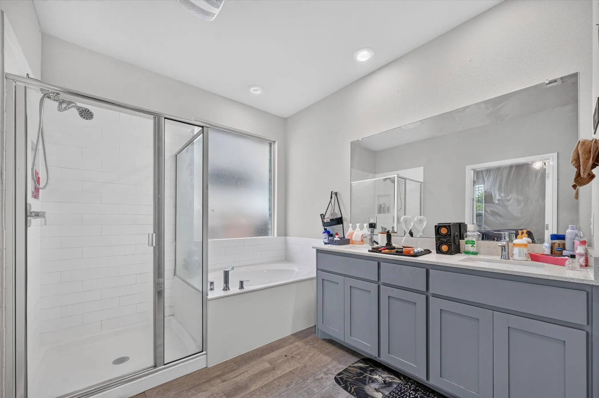 Dual vanity with gray cabinetry and light-toned countertops, large integrated mirror, separate shower with subway tile surround, soaking tub with a frosted window, and wood-finish flooring